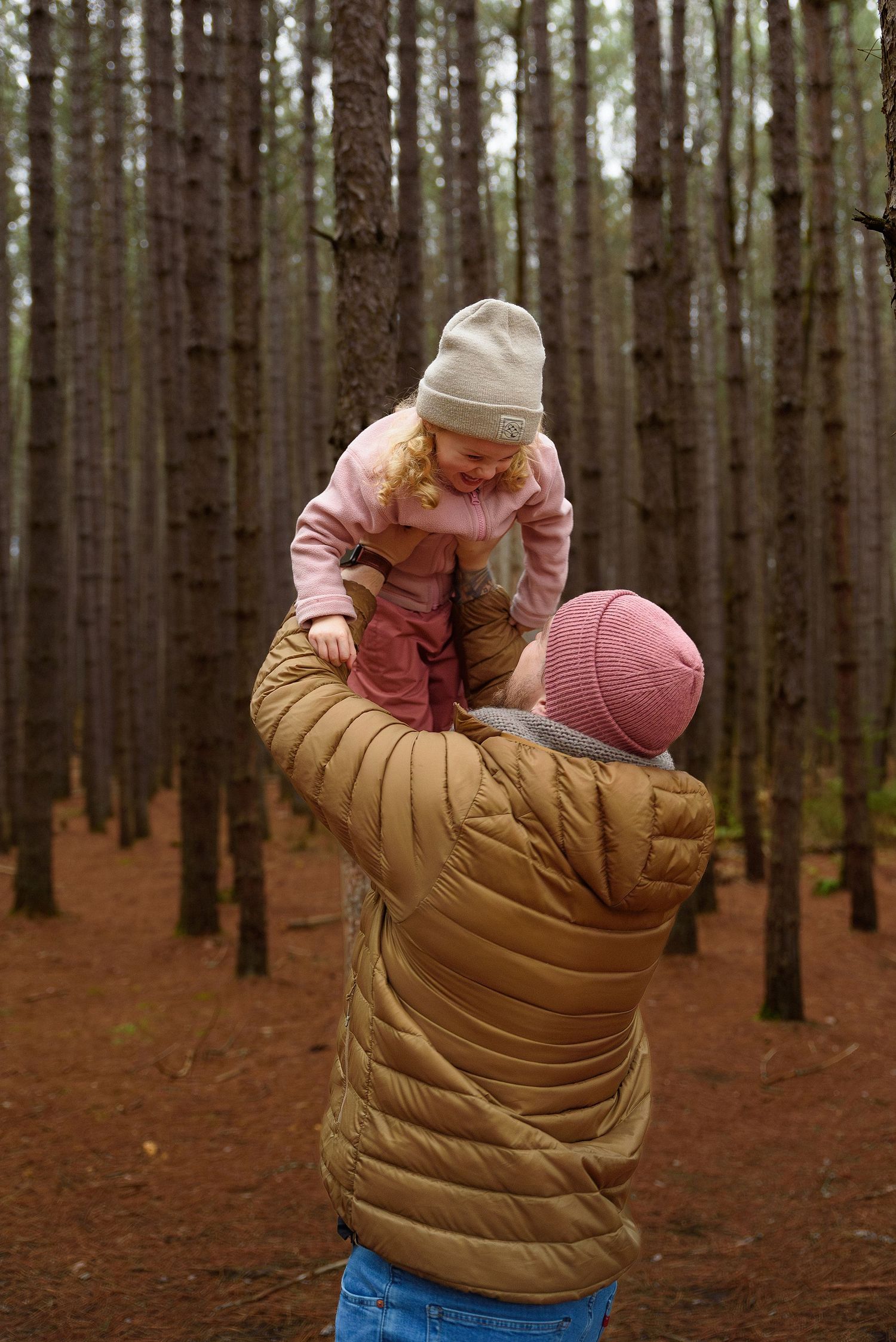 Adult in tan puffer jacket lifting smiling toddler girl in pink coat and hat in a pine forest.