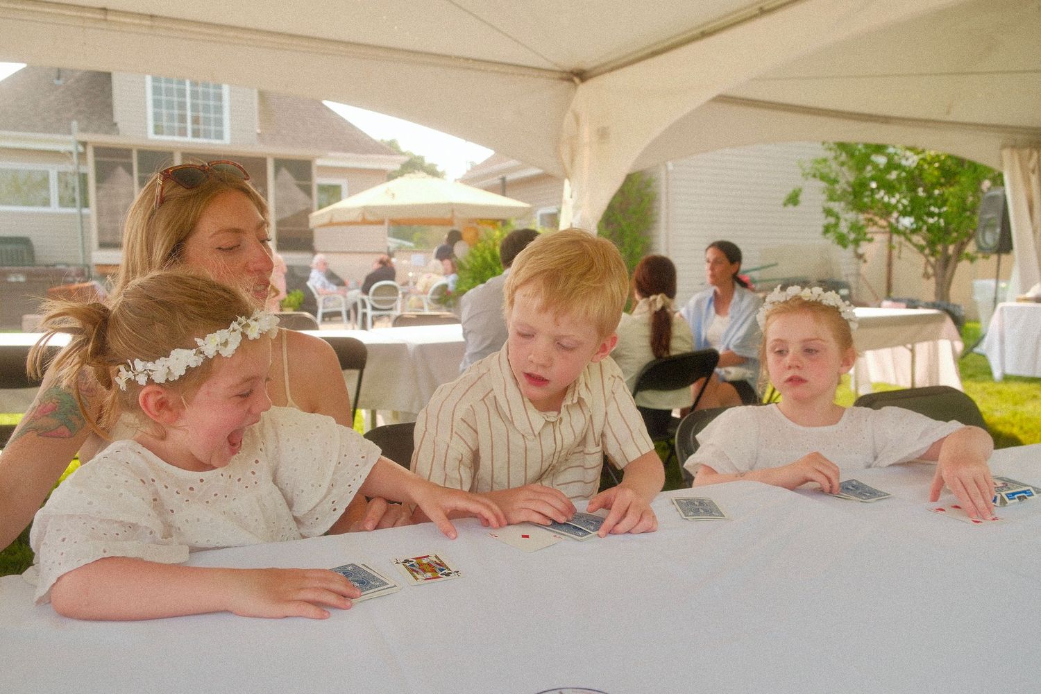 Three young children and a woman sit at a white round table under a tent, looking at cards at an outdoor event.