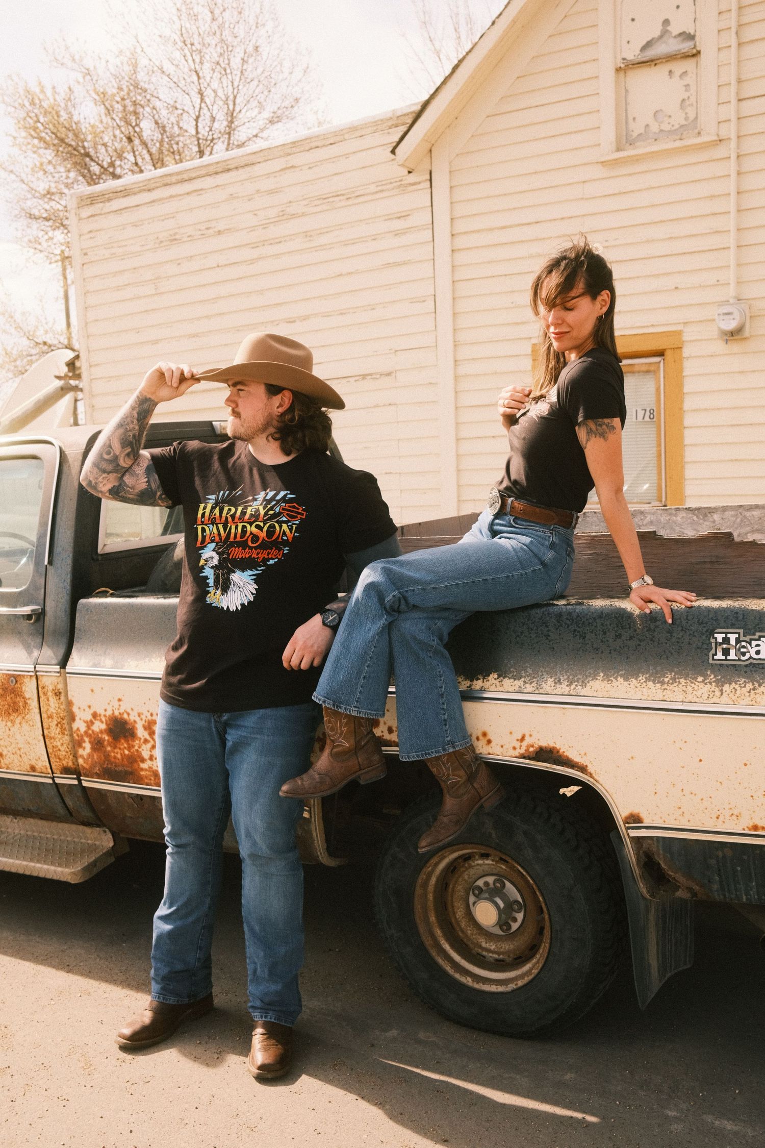 Two women in Harley-Davidson tees, jeans, and boots pose by a rusty vintage truck outside a rural building.