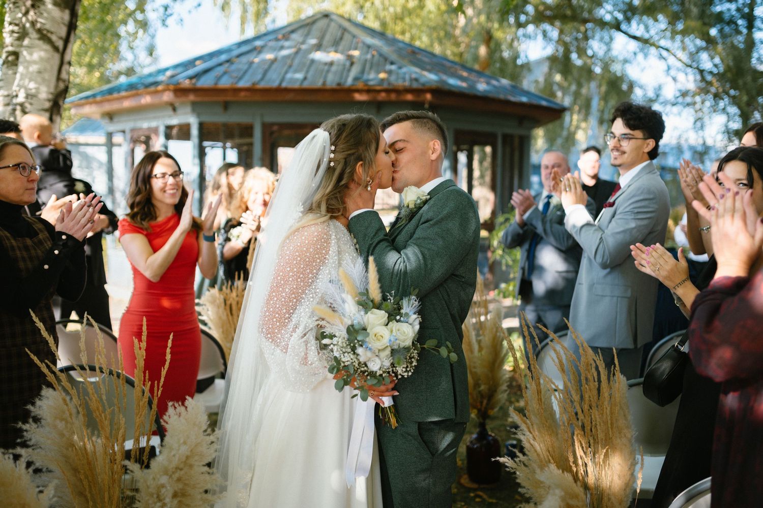 Wedding ceremony guests gathered outdoors under trees with decorative pampas grass.
