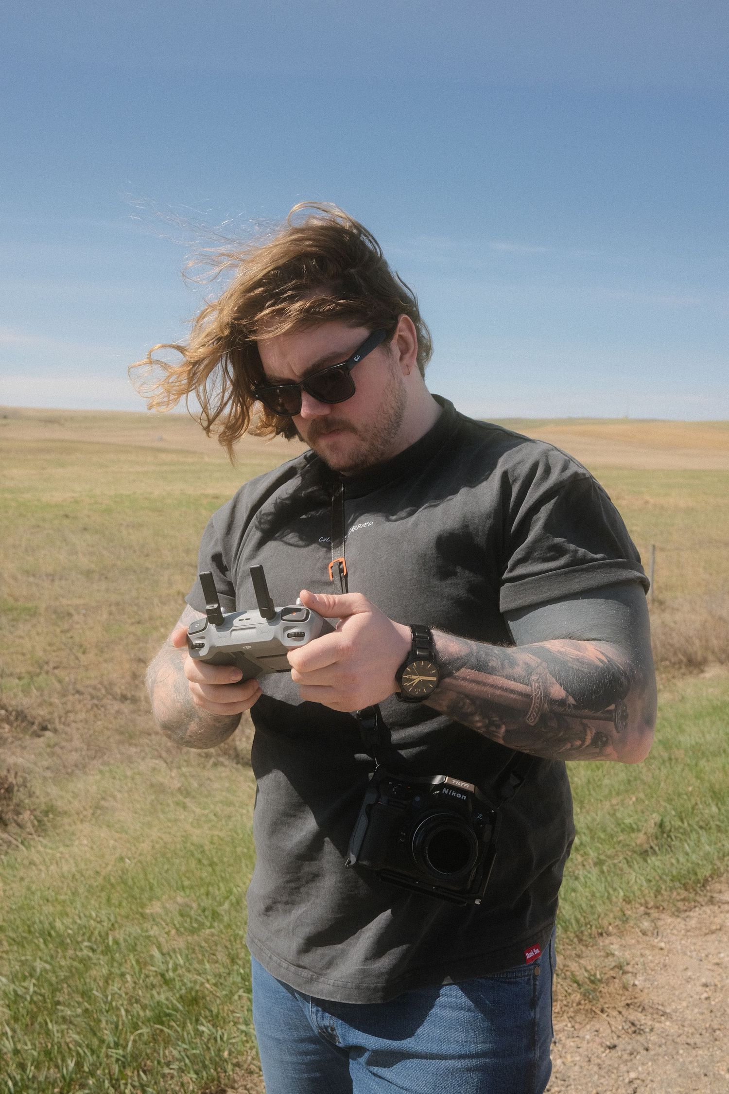 Tattooed man with long hair and sunglasses operating a drone remote controller in an open grassy field.