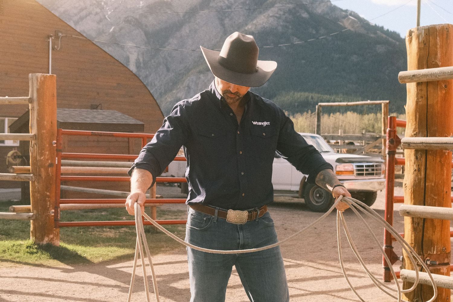 Cowboy in black hat and Wrangler shirt handling rope at wooden corral with mountain backdrop.