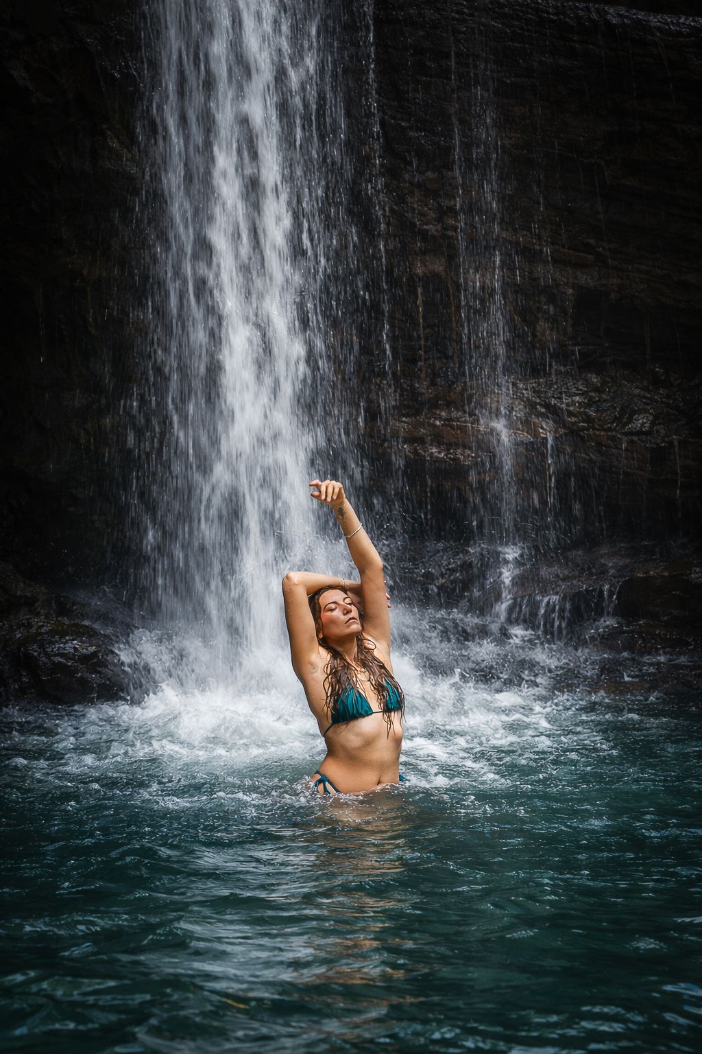 A person enjoys swimming and posing in turquoise waters beneath a cascading waterfall in a tropical paradise.