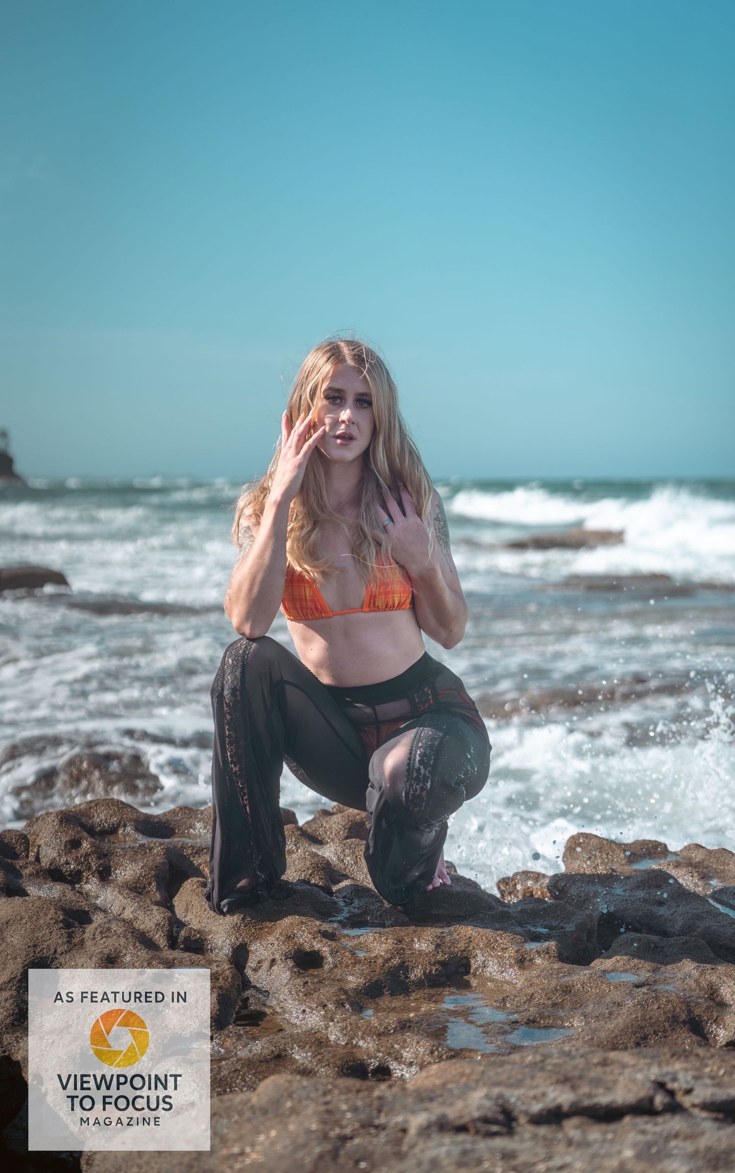 Model poses on rocky beach with crashing waves and blue sky in dramatic fashion photoshoot series.