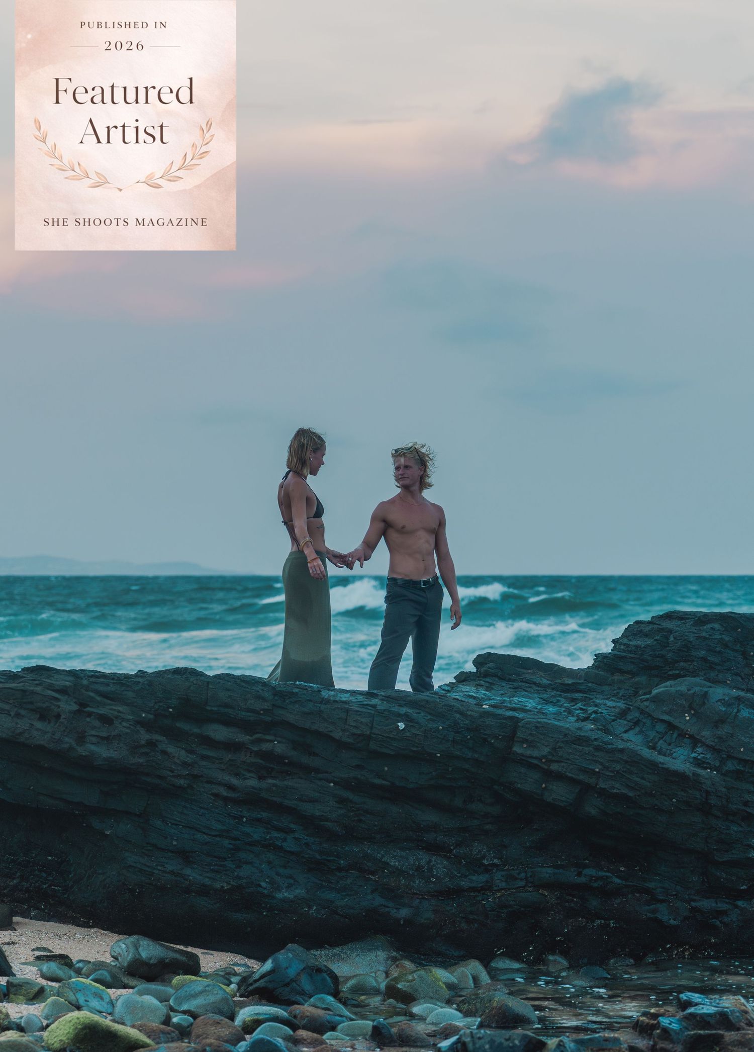 Couple holding hands on rocky shoreline at dusk, featured in She Shoots Magazine 2026 as a Featured Artist.