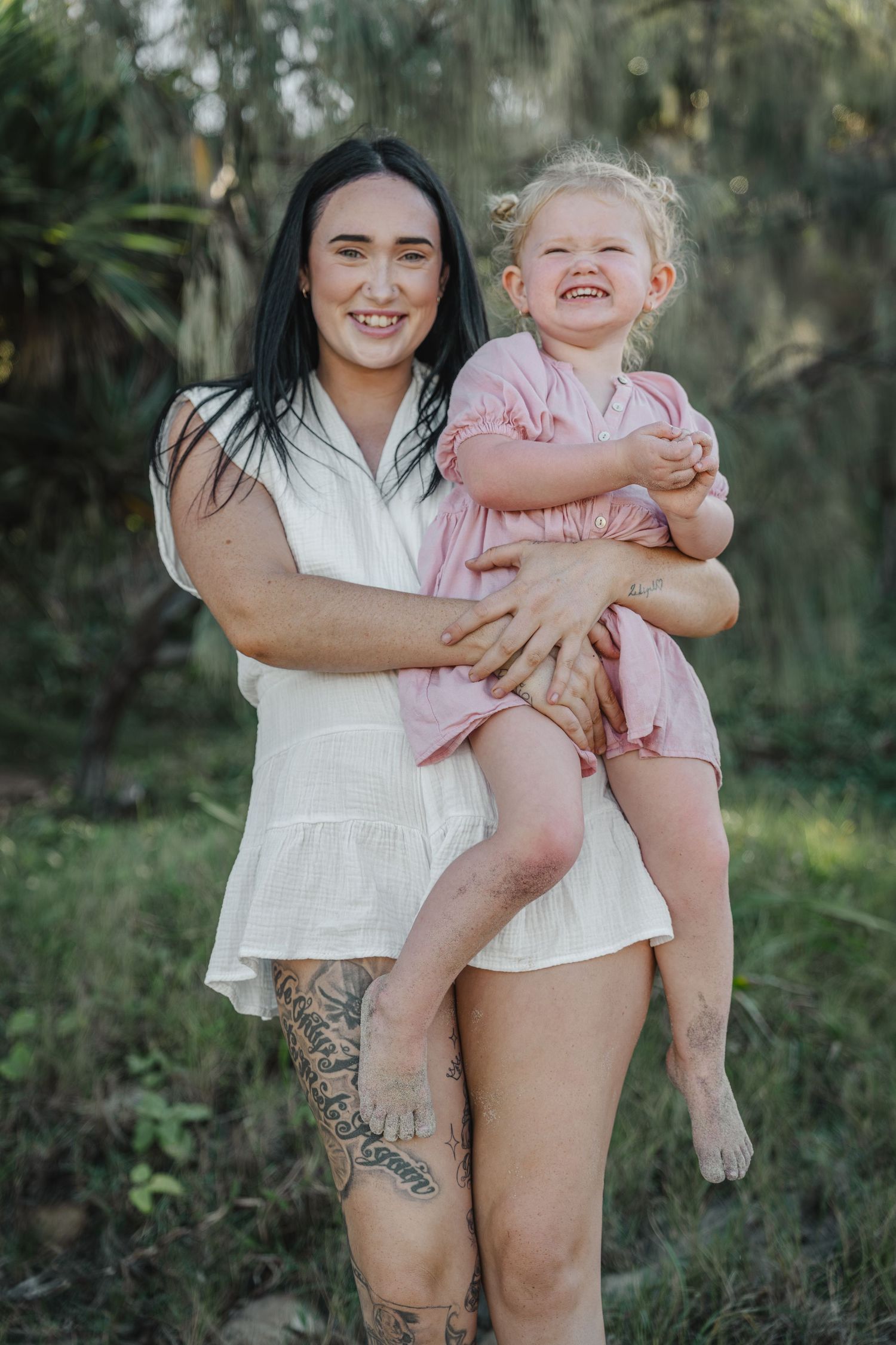 A smiling woman with dark hair and tattoos holds a laughing toddler girl in a pink dress outdoors.