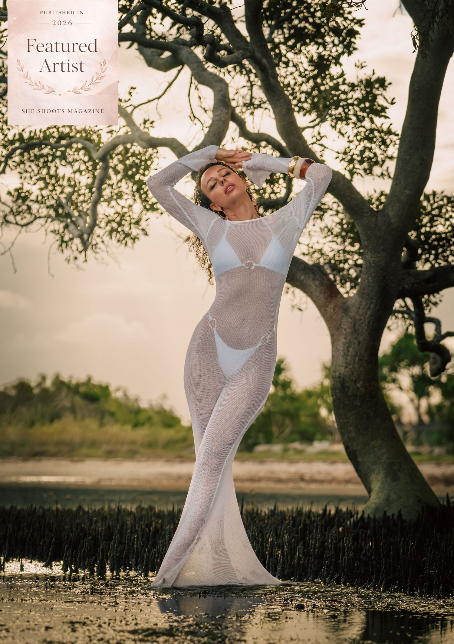 A woman in a sheer white gown poses gracefully outdoors near trees, featured in She Shoots Magazine 2026.
