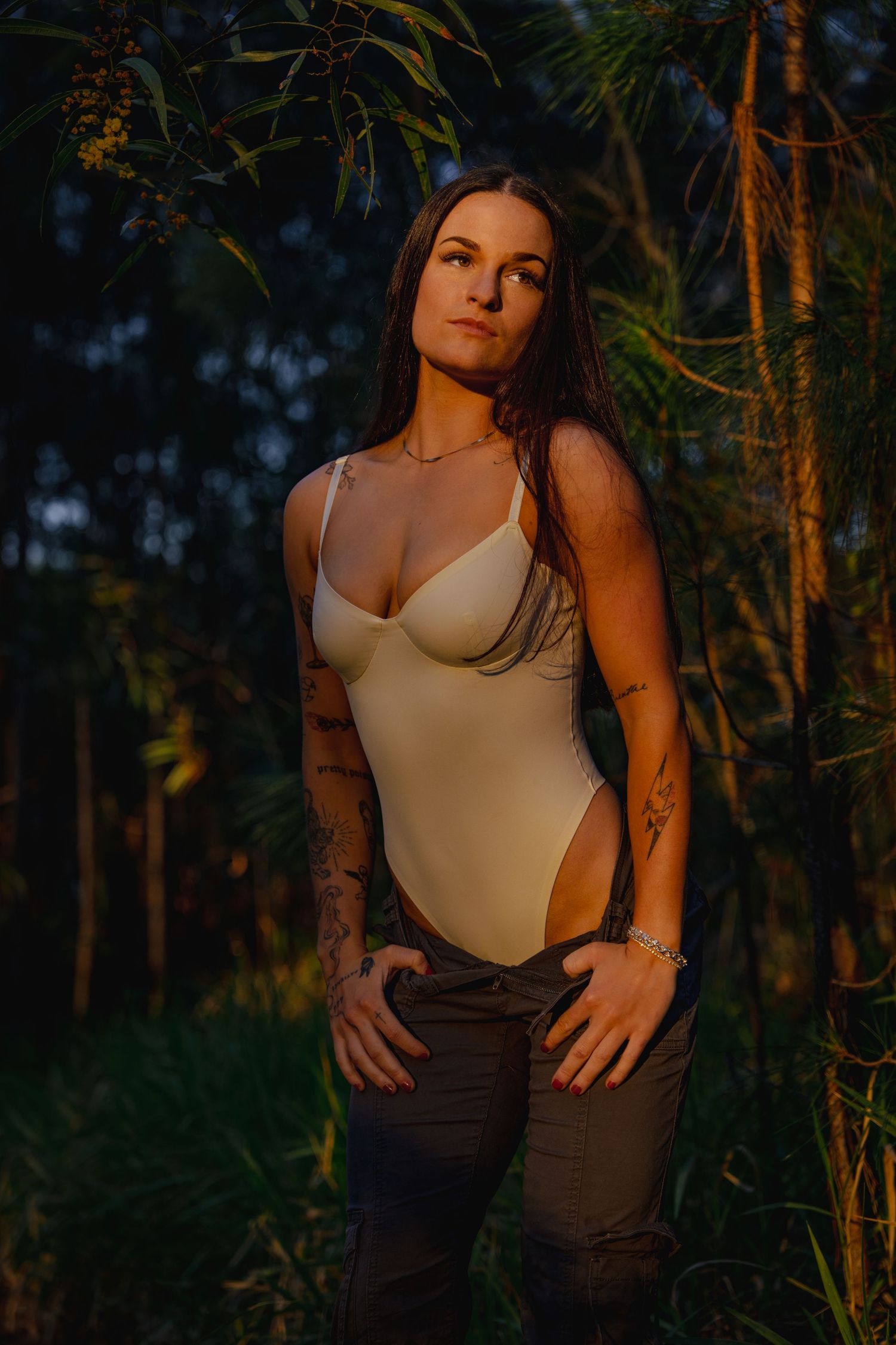Tattooed woman in beige bodysuit and jeans poses outdoors at golden hour surrounded by lush tropical greenery.