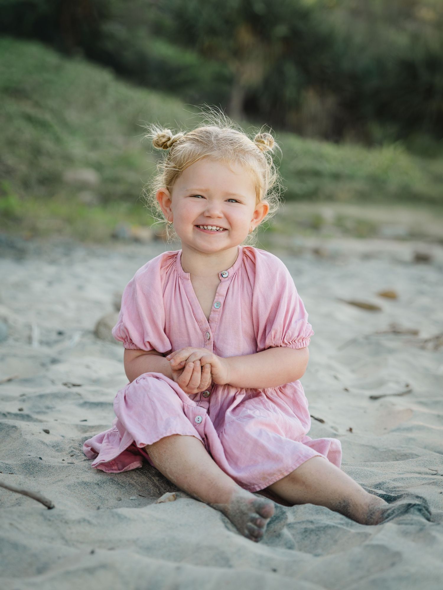 A smiling toddler girl in a pink dress sits barefoot on sandy beach with hair in two buns.