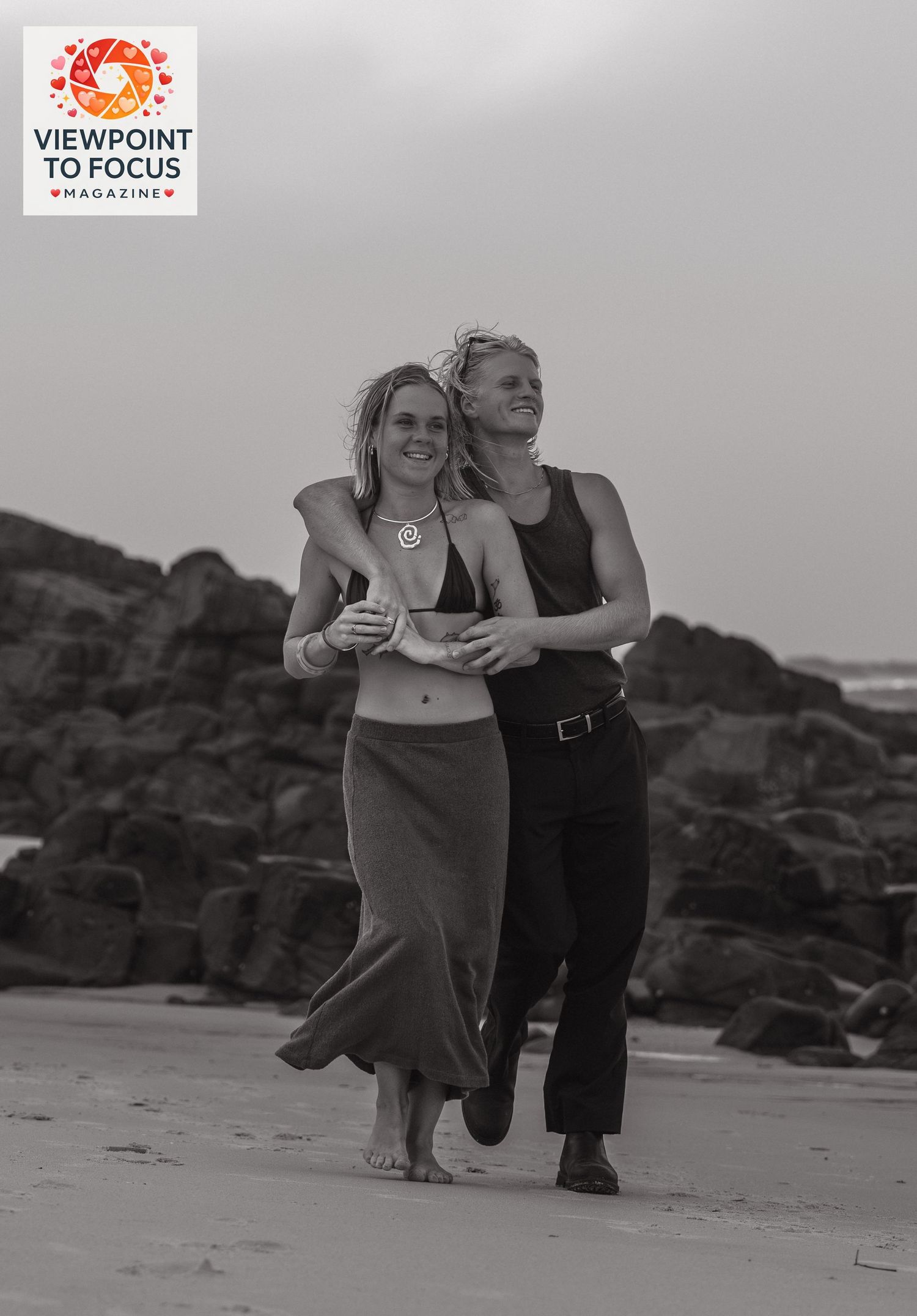 Two women walk arm in arm on a rocky beach in a black and white photo, one in a bikini top and skirt.