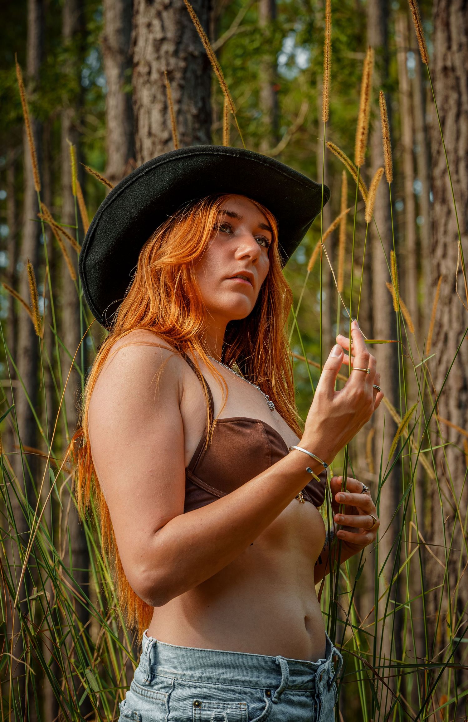 A person in a black cowboy hat and brown top stands among tall reeds in a forest setting at golden hour.