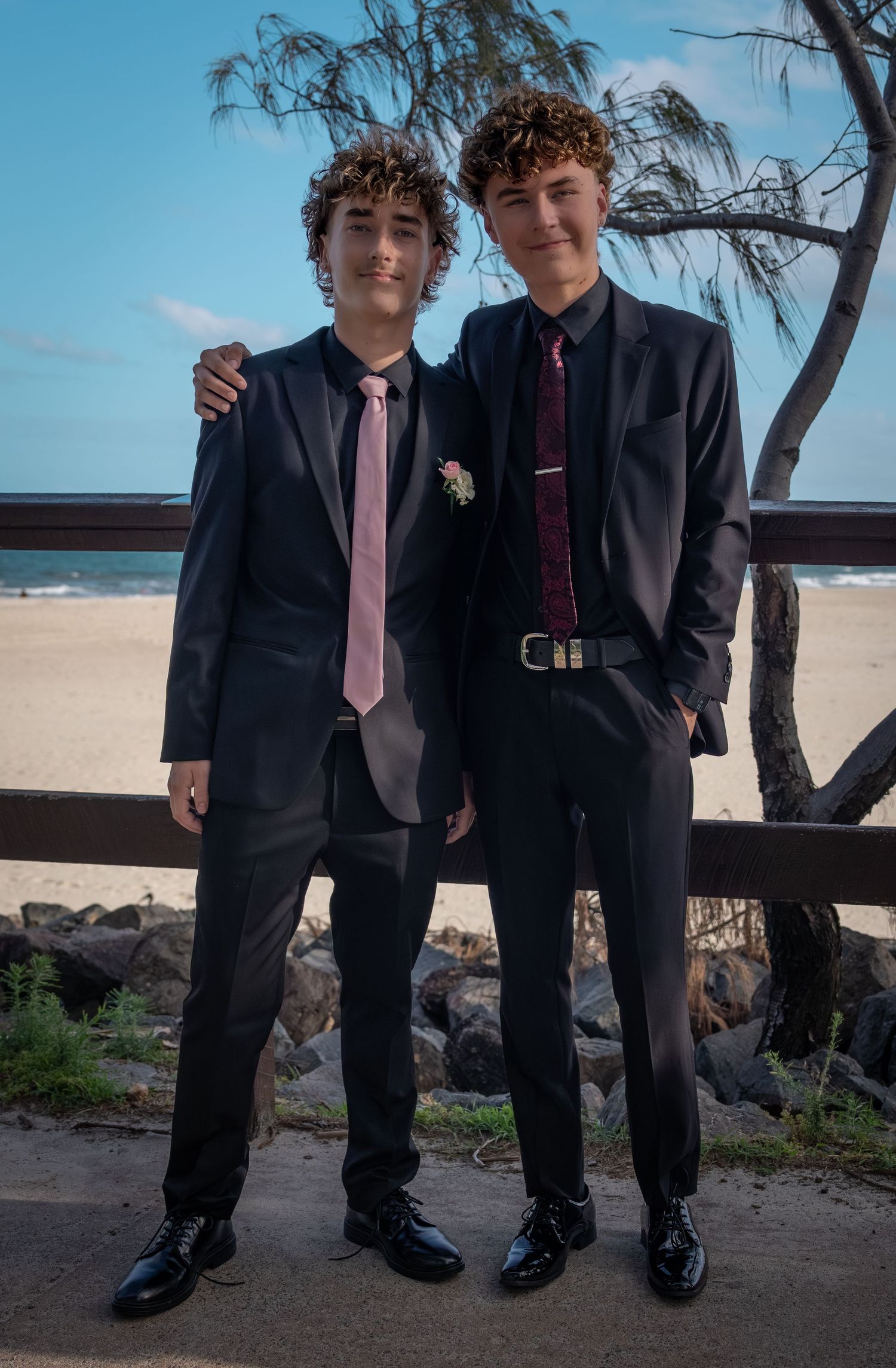 Two young people in formal black suits and ties standing on a beach.