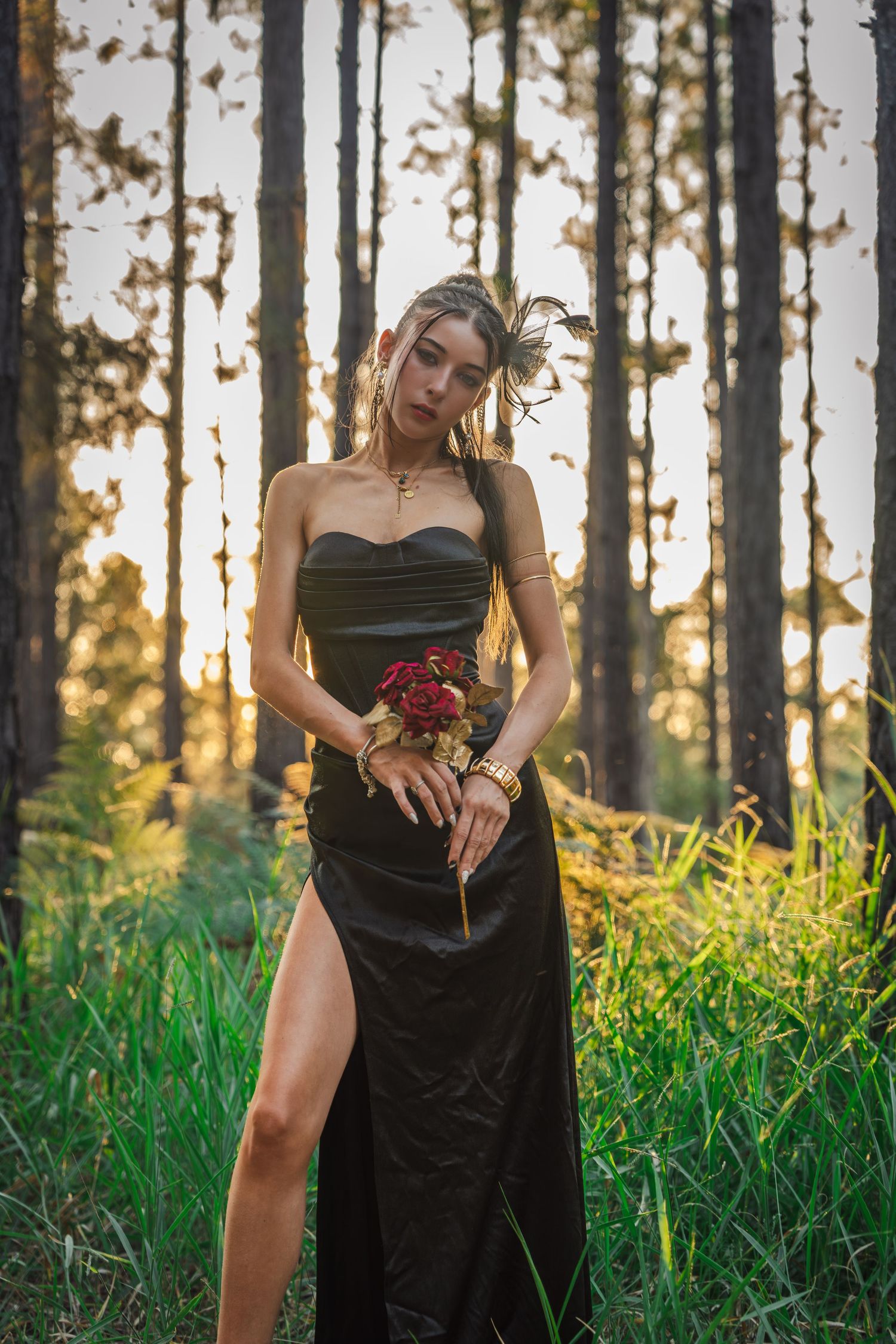 Woman in black strapless gown holding red roses in a sunlit forest at golden hour.