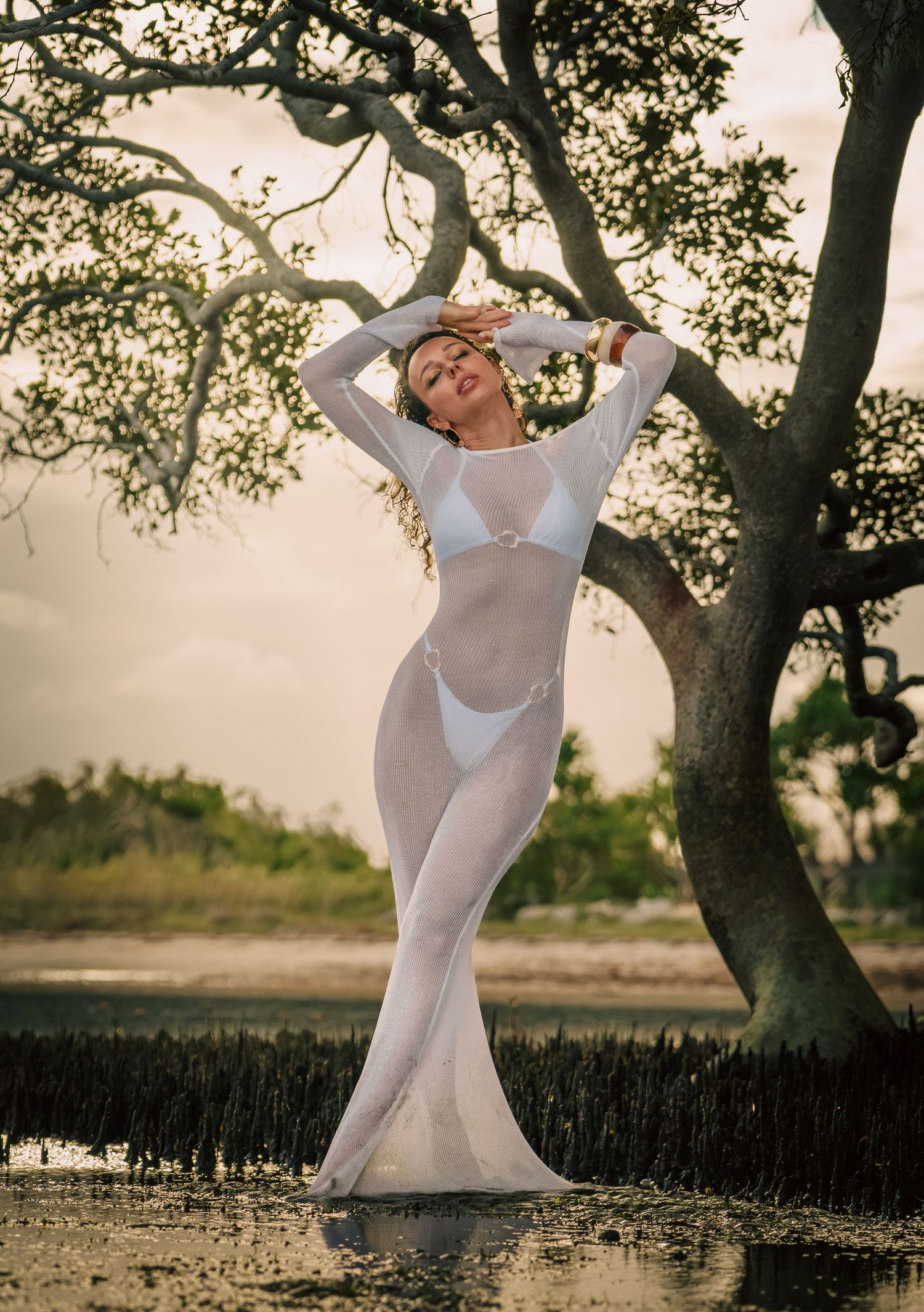 Woman in white sheer mesh dress poses gracefully near trees by water at sunset.