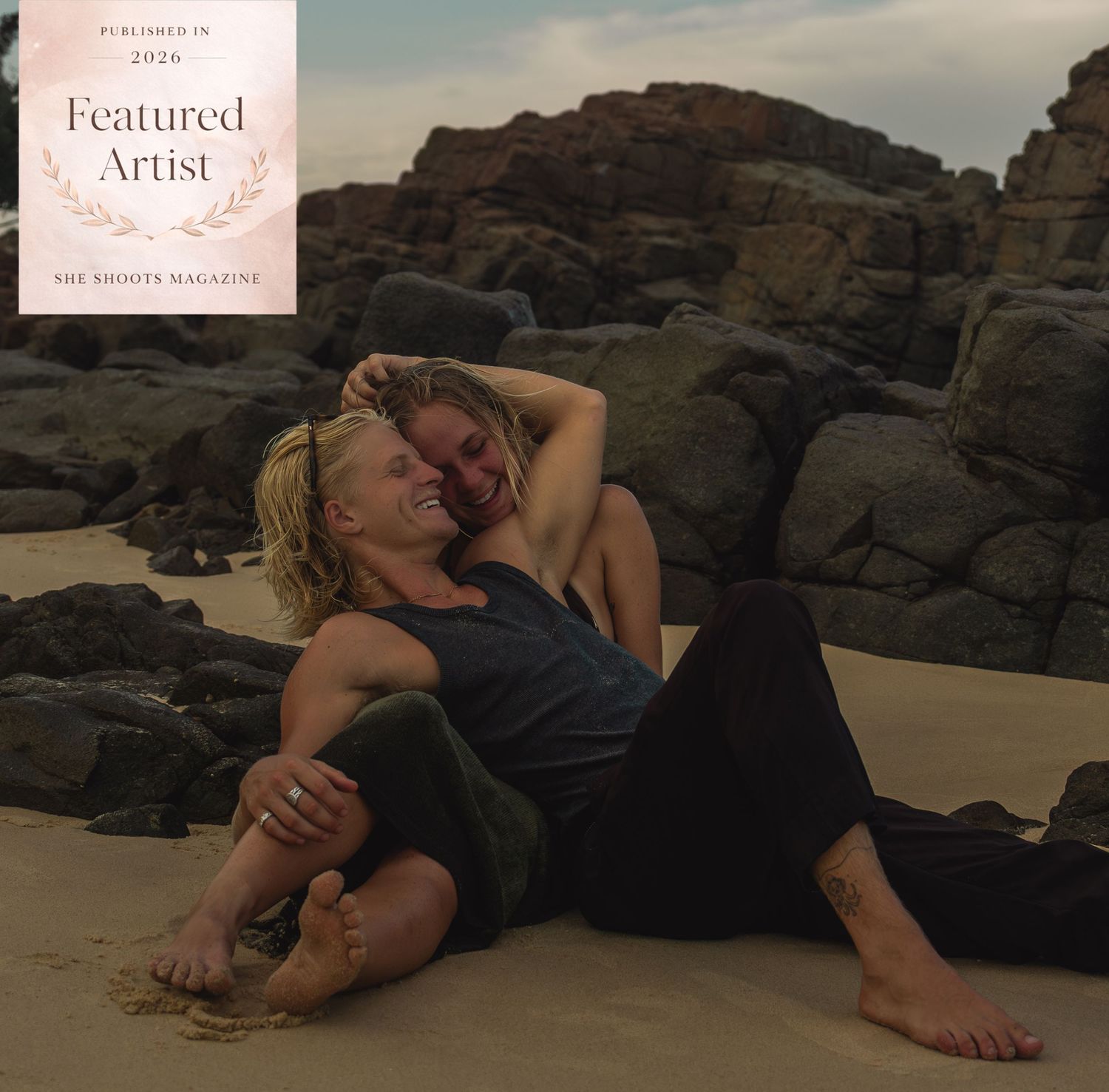 Two people embrace romantically on a sandy beach with rocky cliffs, featured in She Shoots Magazine 2026.