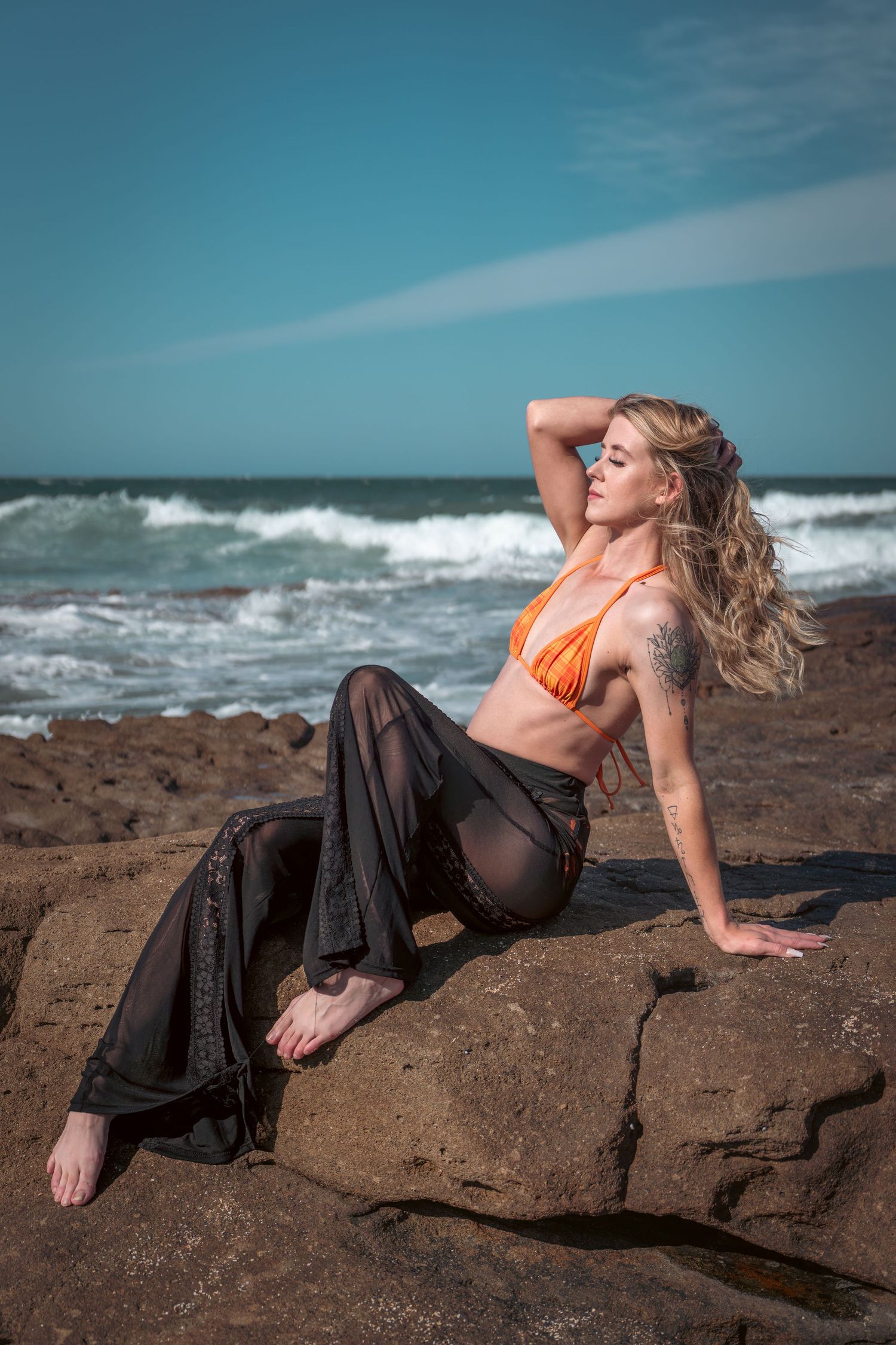 Blonde woman in orange bikini top and sheer black pants posing on rocky beach with ocean waves behind her.