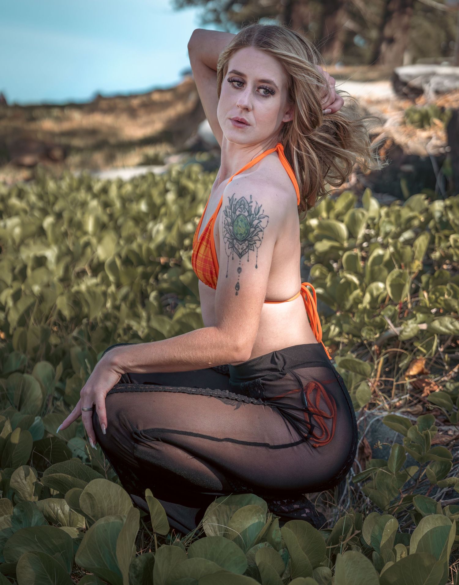 Fashion portrait in orange top and sheer pants amid coastal vegetation.