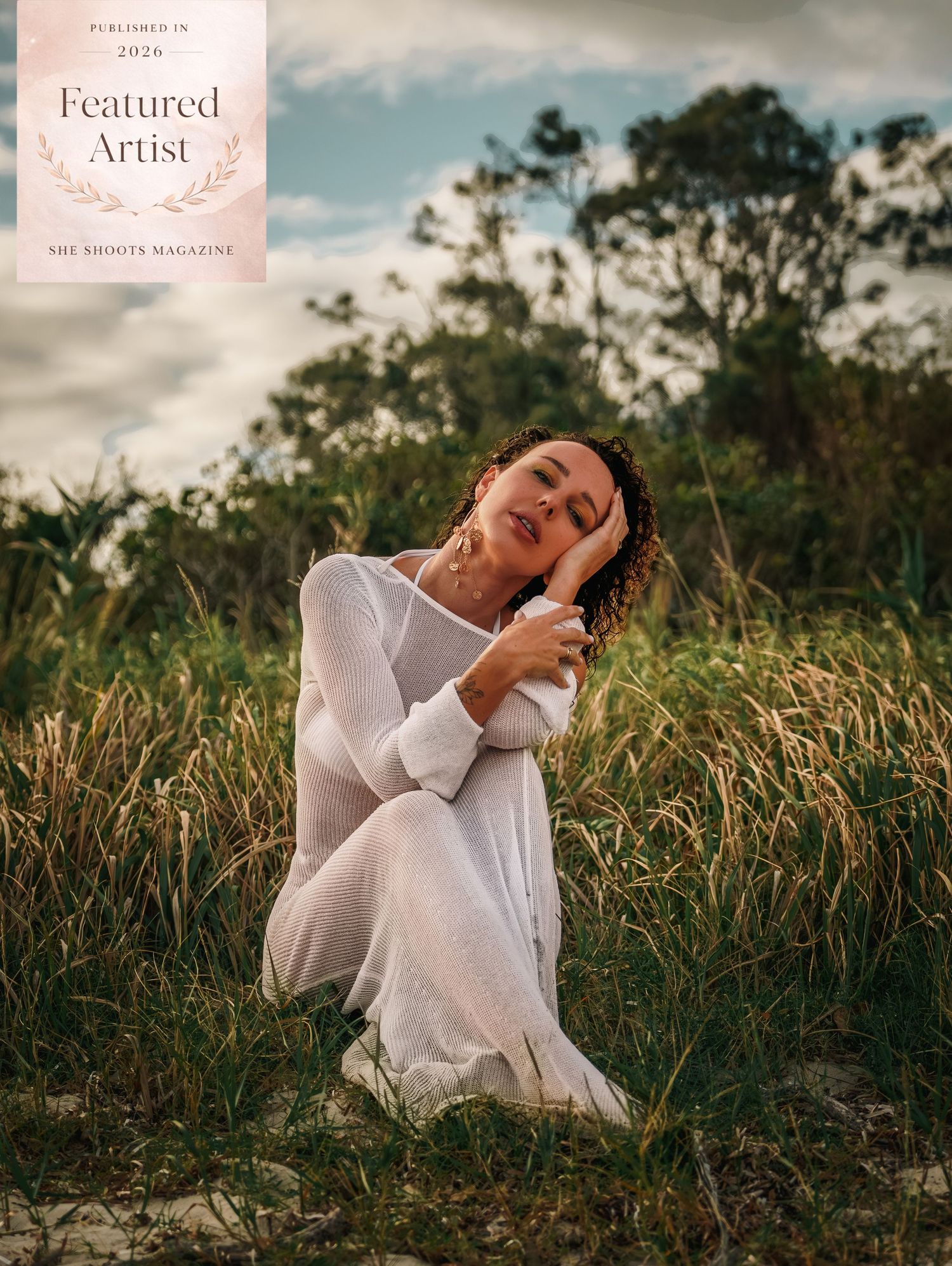 Woman in white dress kneeling in tall grass field, featured artist cover for She Shoots Magazine 2026.