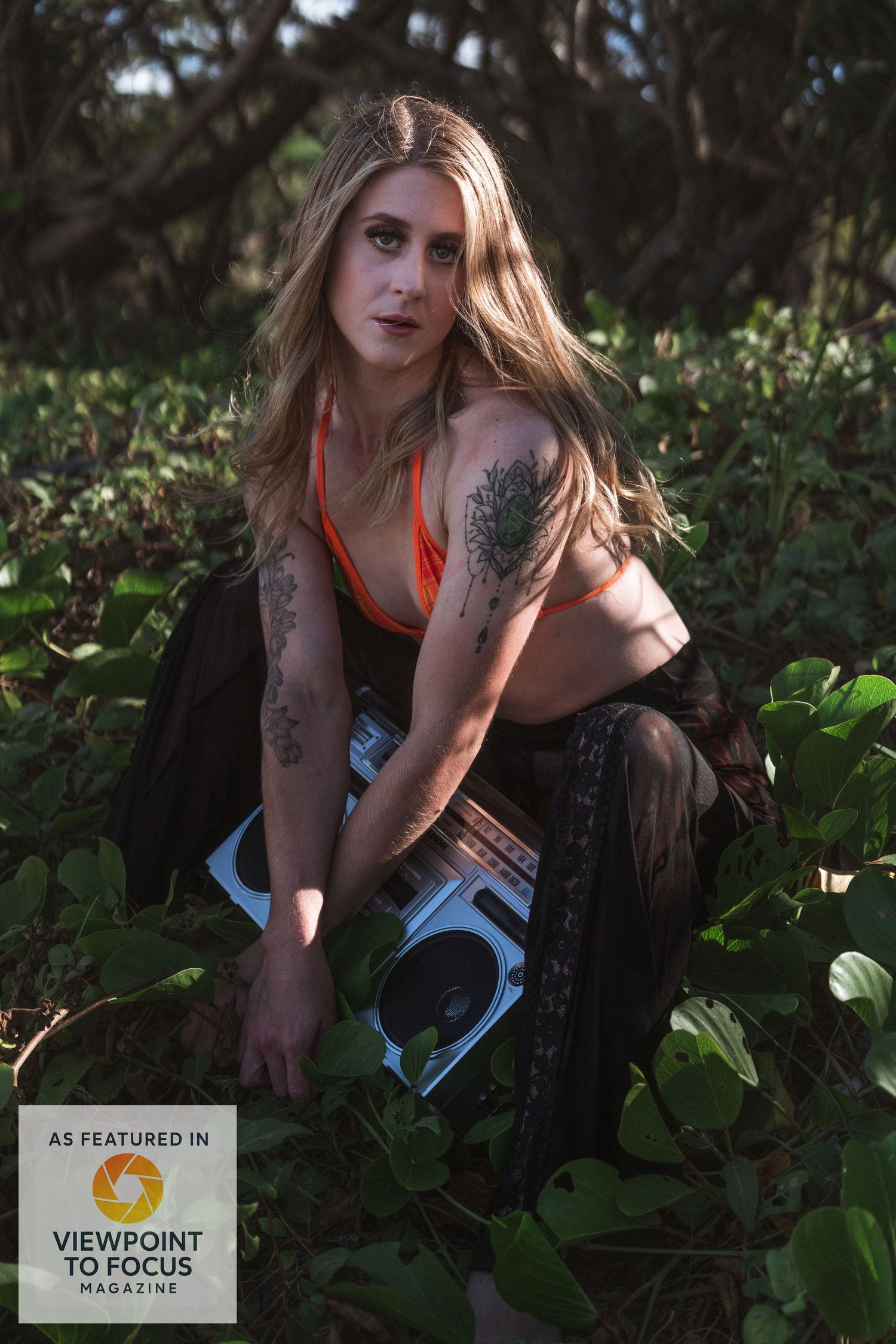 A model in orange athletic wear poses in a lush forest setting with dappled sunlight filtering through the trees.