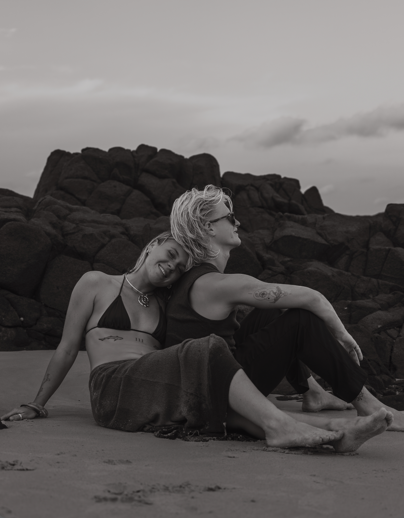 Couple sitting together on a rocky beach in black and white, one leaning on the other, both barefoot at sunset.