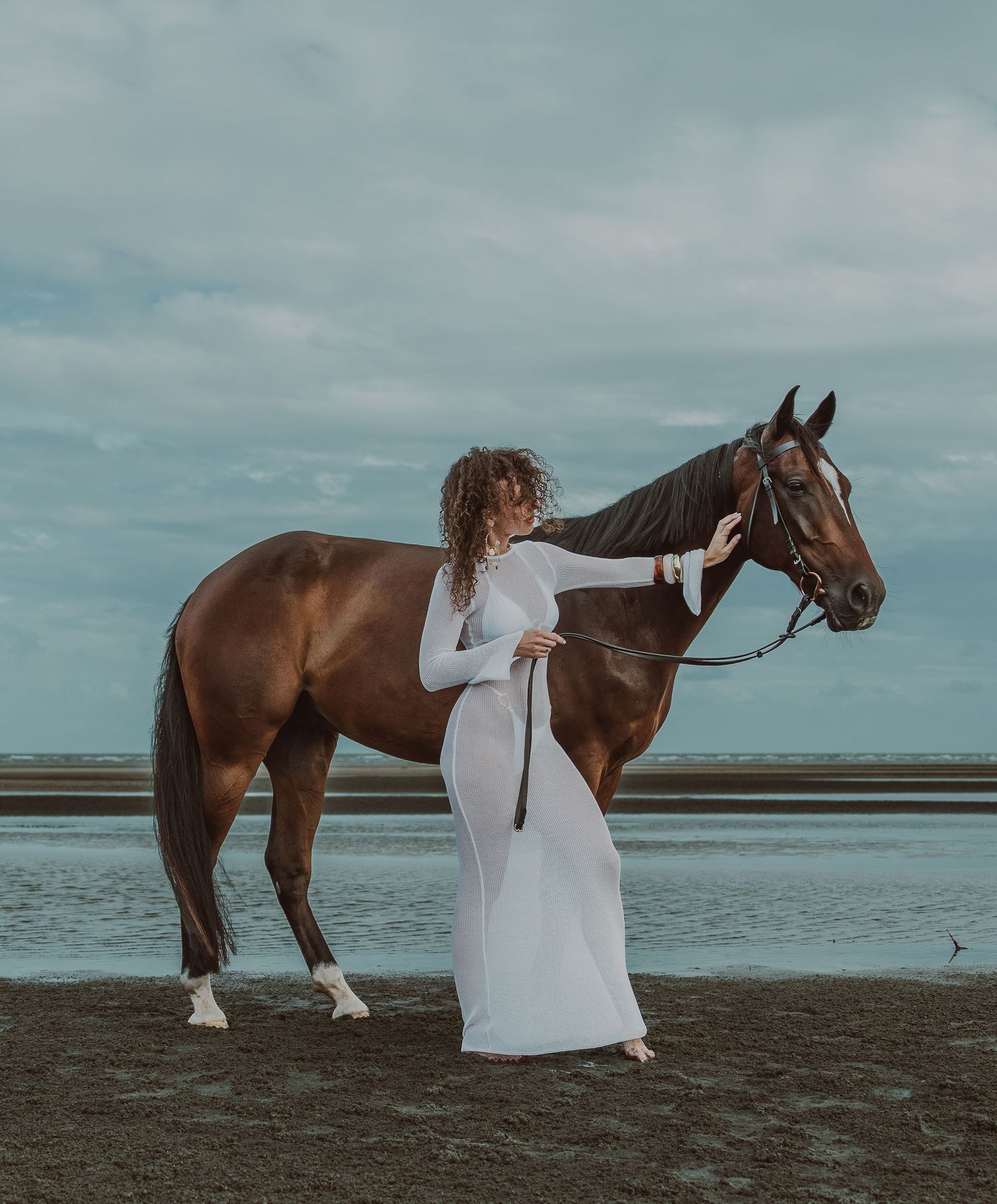 Woman in white dress standing beside a brown horse on a beach with cloudy sky backdrop.