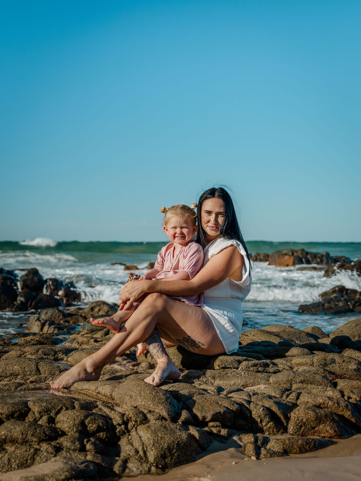 A young mother sits on rocky beach holding her toddler daughter, with ocean waves and clear blue sky behind them.
