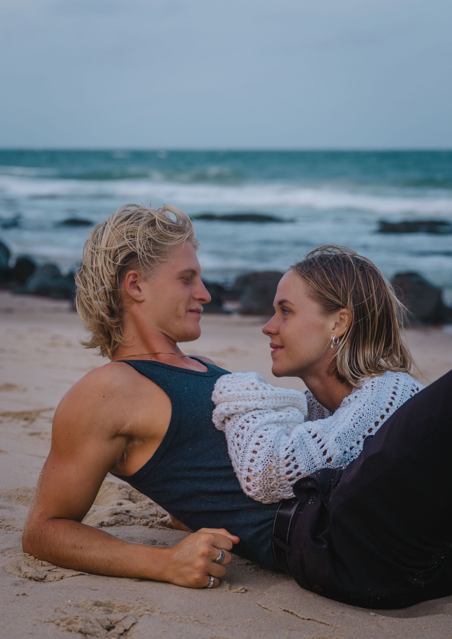 A couple lying on a beach at sunset sharing intimate moments with ocean waves in the background.