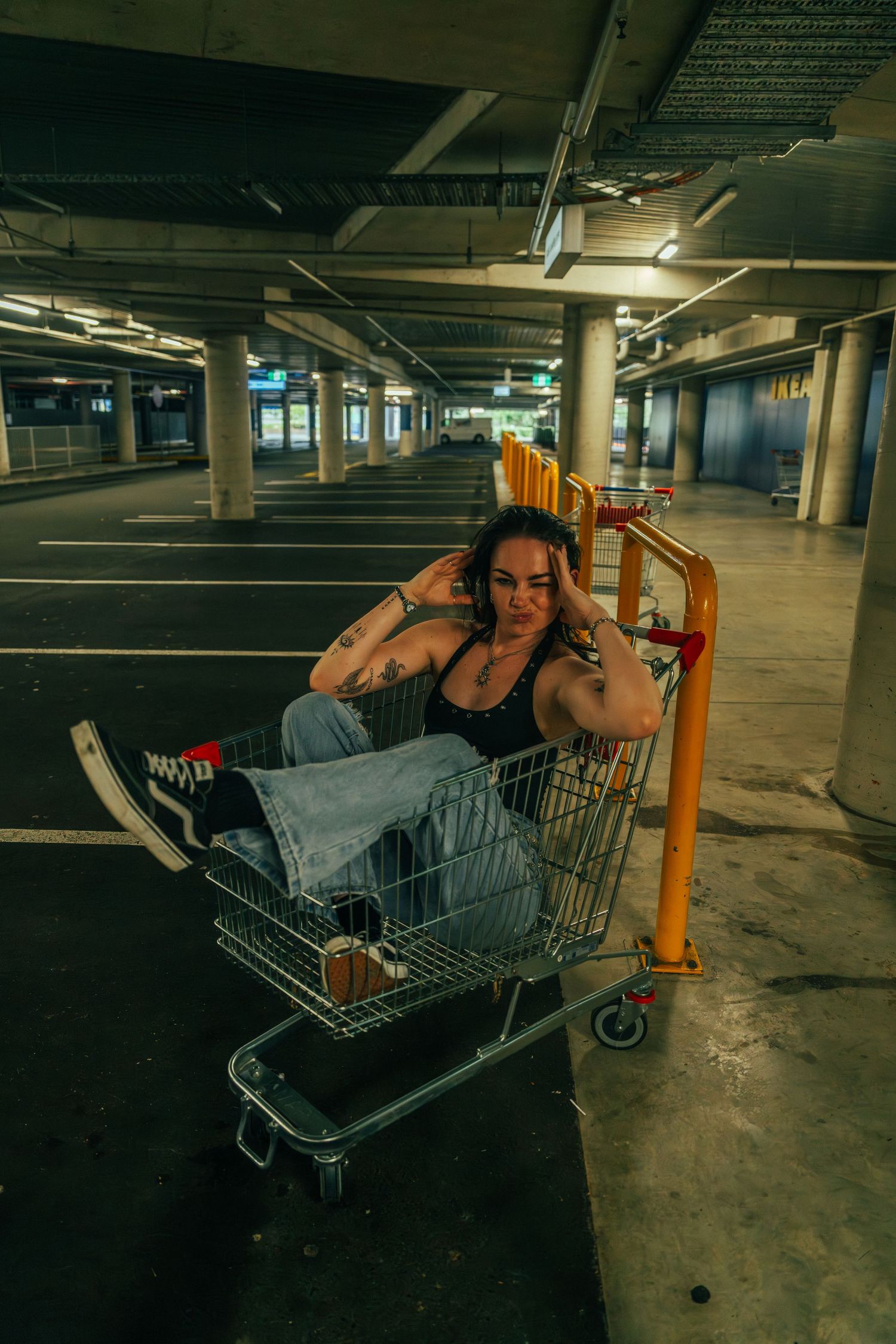A tattooed woman sits inside a shopping cart in a dimly lit parking garage, wearing jeans and a black tank top.