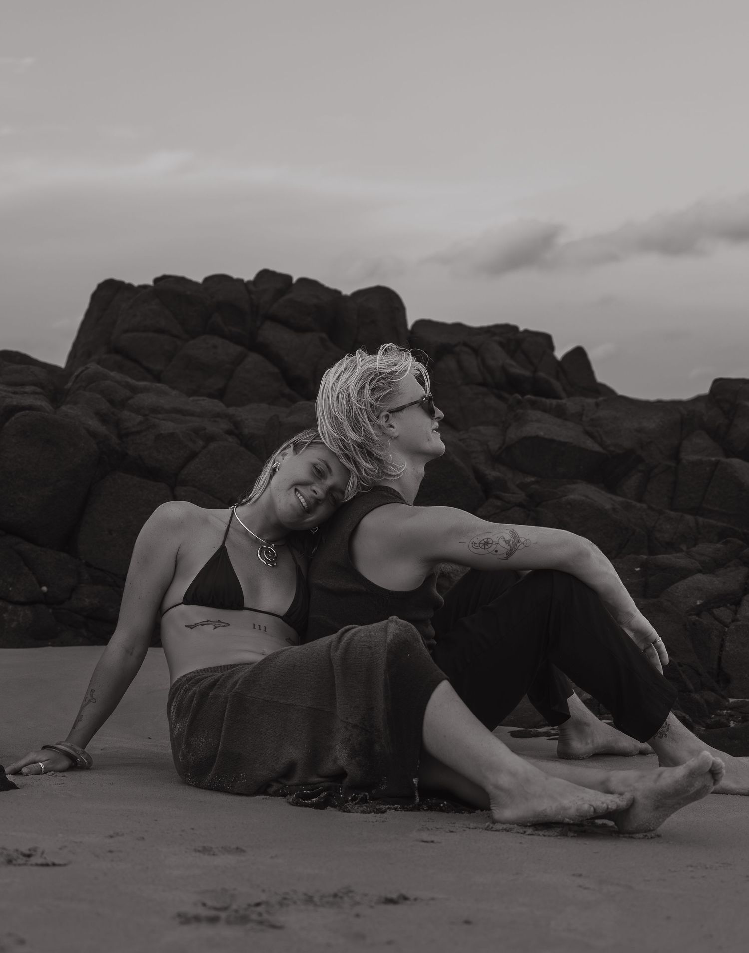 Two people in casual attire sitting on a sandy beach with dramatic rock formations in the background.