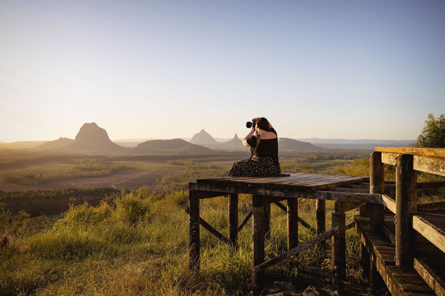 Photographer sitting on wooden lookout platform capturing stunning mountain landscape at golden hour sunset.