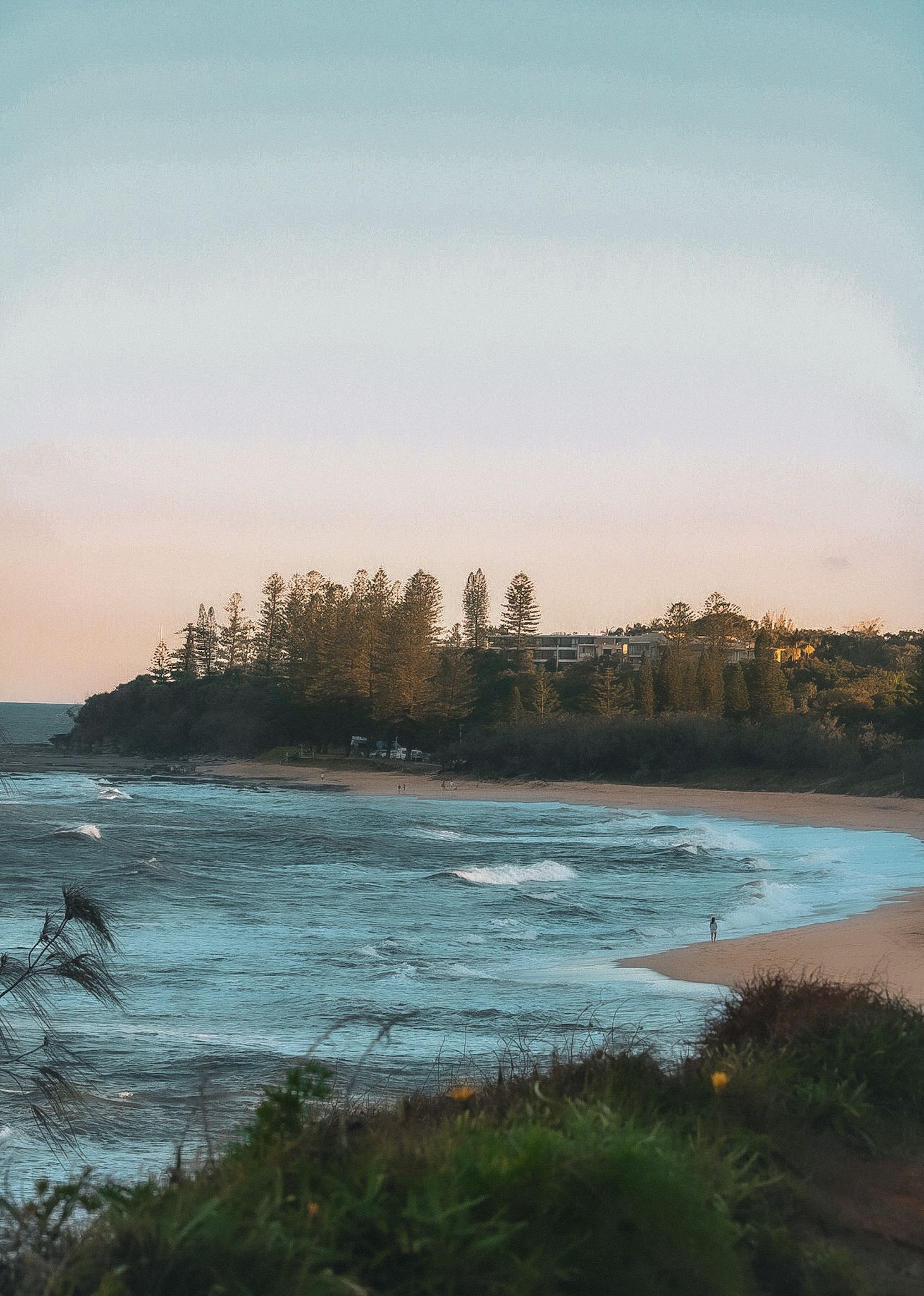 Coastal beach scene at dusk with rolling waves, sandy shore, lush headland trees, and a lone figure near the water.