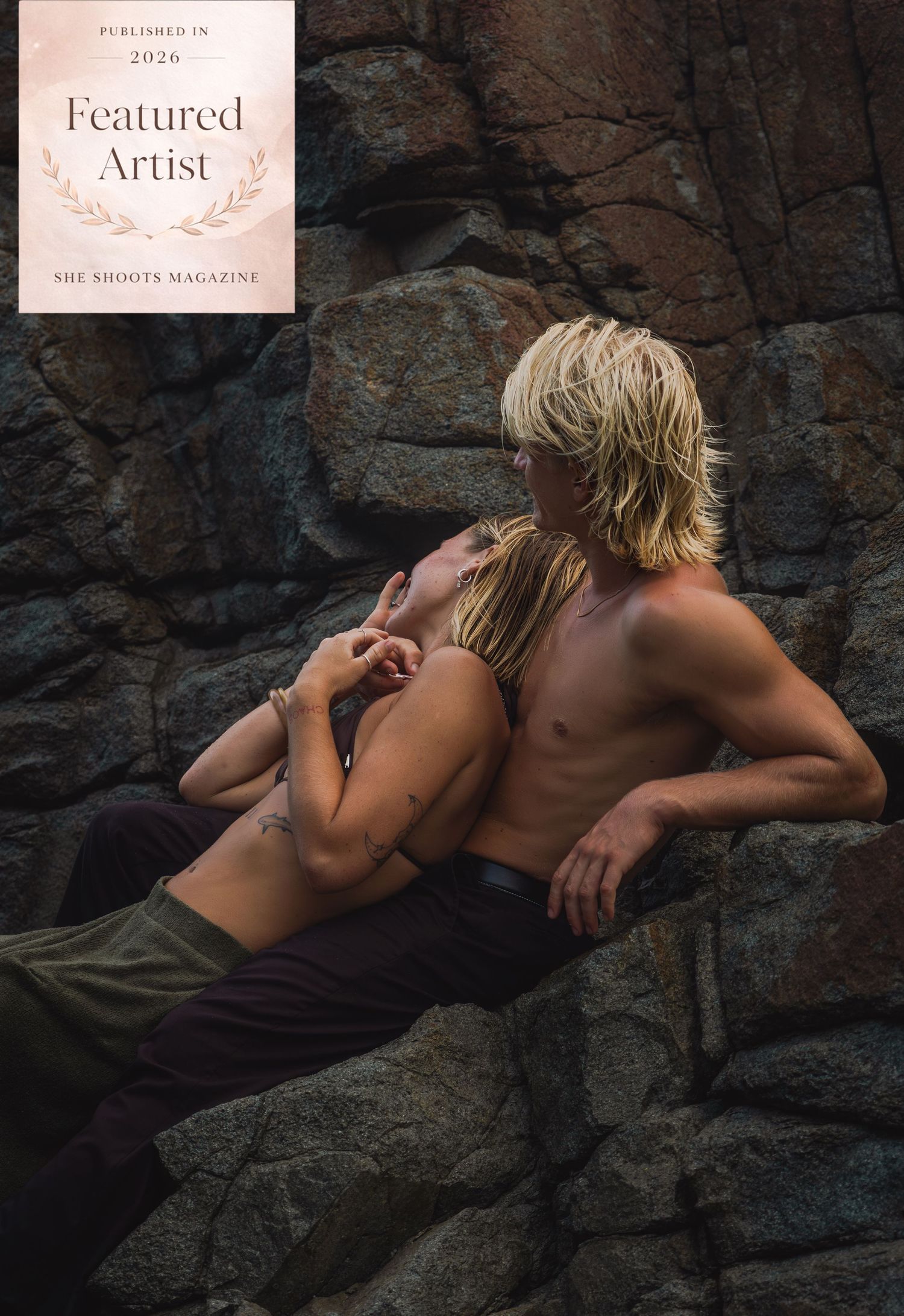 A shirtless blonde man and woman embrace romantically against rugged dark rocks, featured in She Shoots Magazine 2026.
