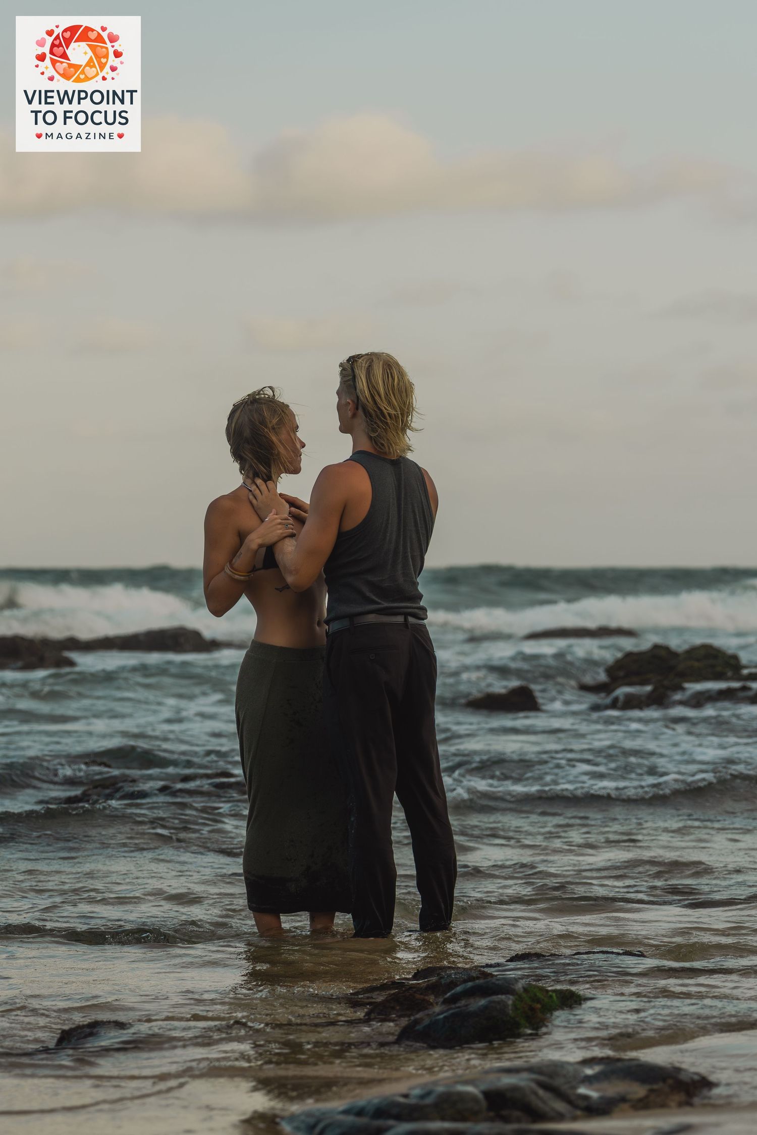 Couple embracing romantically at ocean shore, standing in shallow waves on rocky beach at dusk.