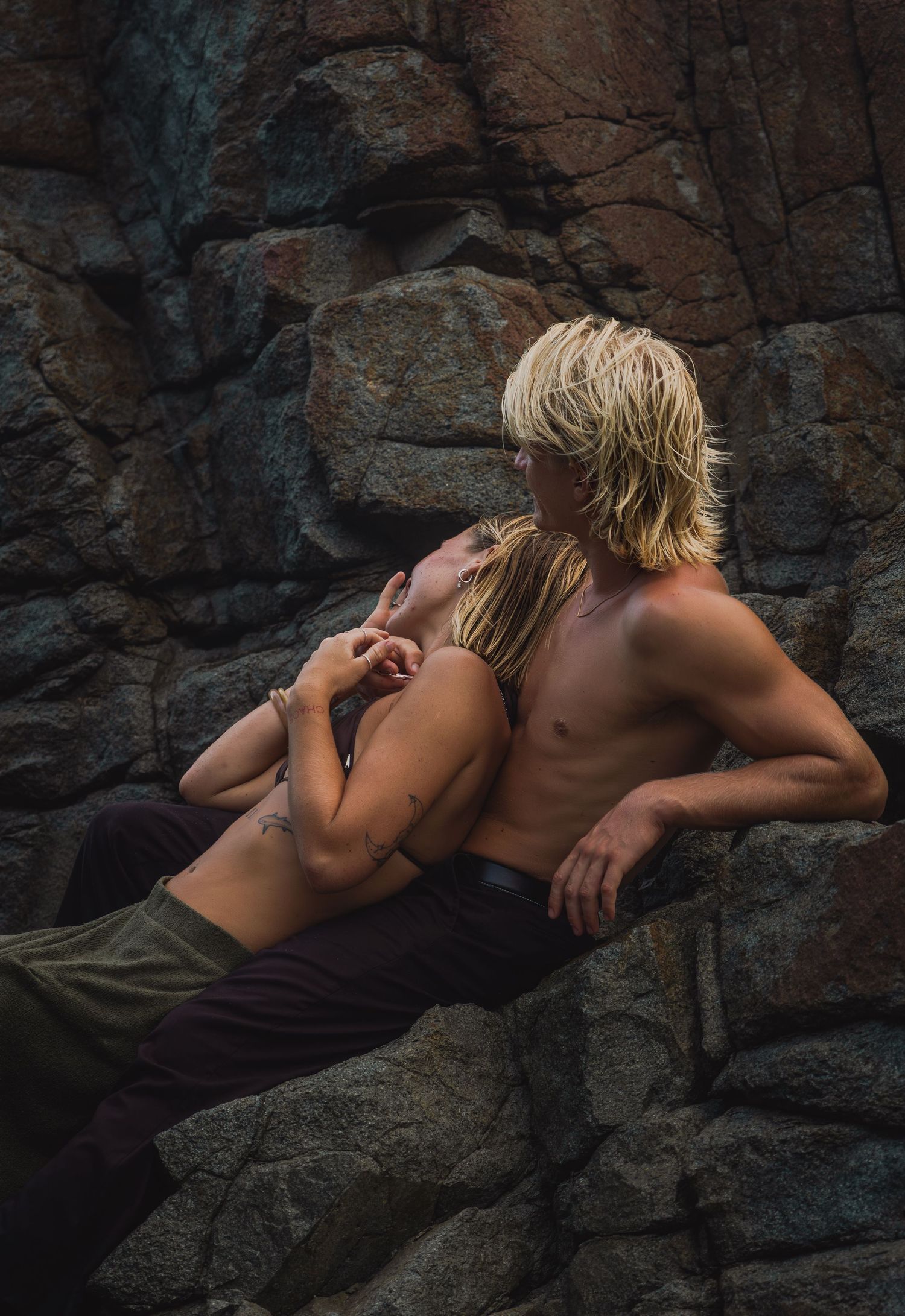 Two blond individuals embrace romantically against a rugged rocky cliff backdrop outdoors.
