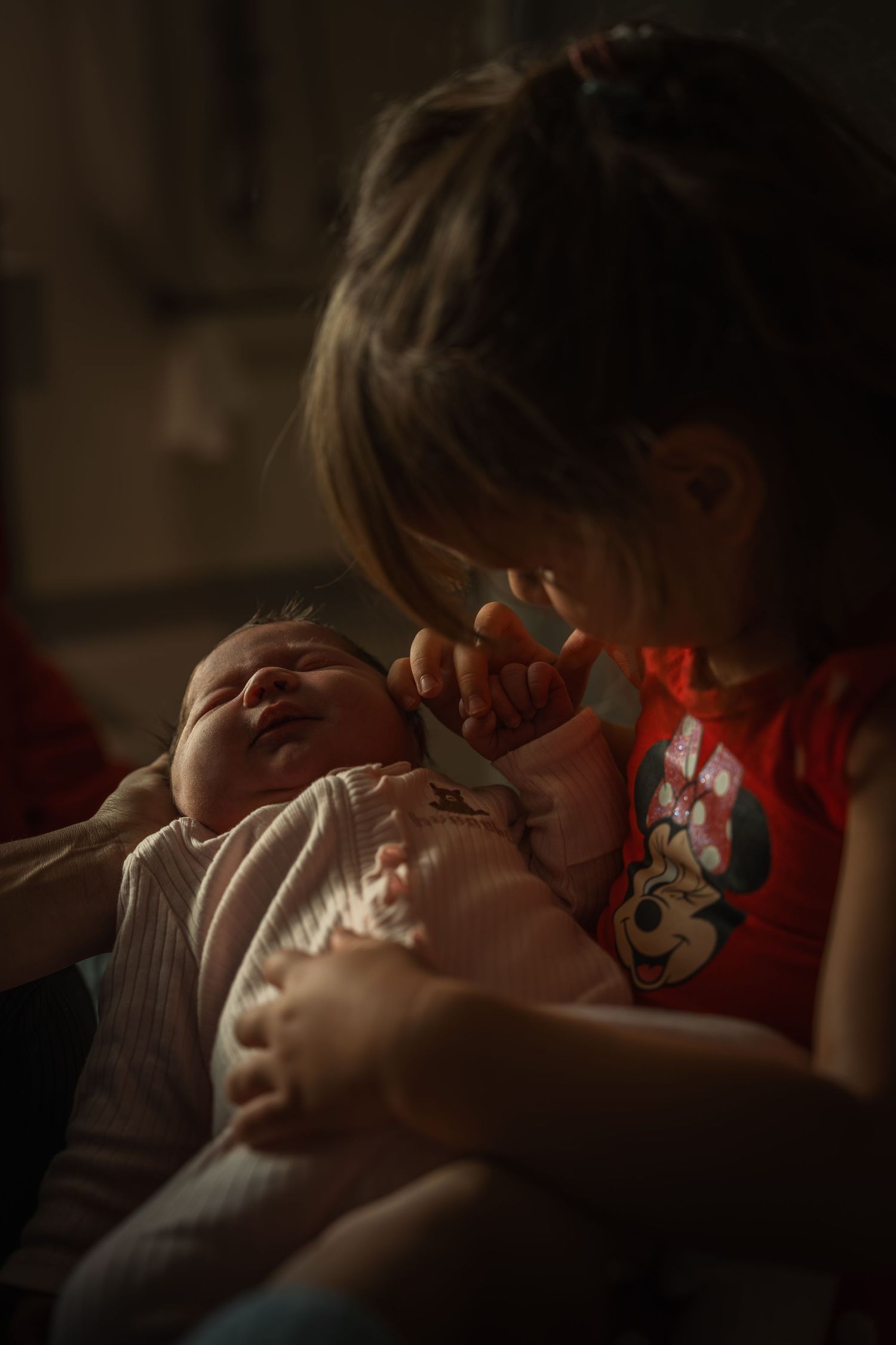A sleeping infant in a white onesie being held tenderly in dim lighting.