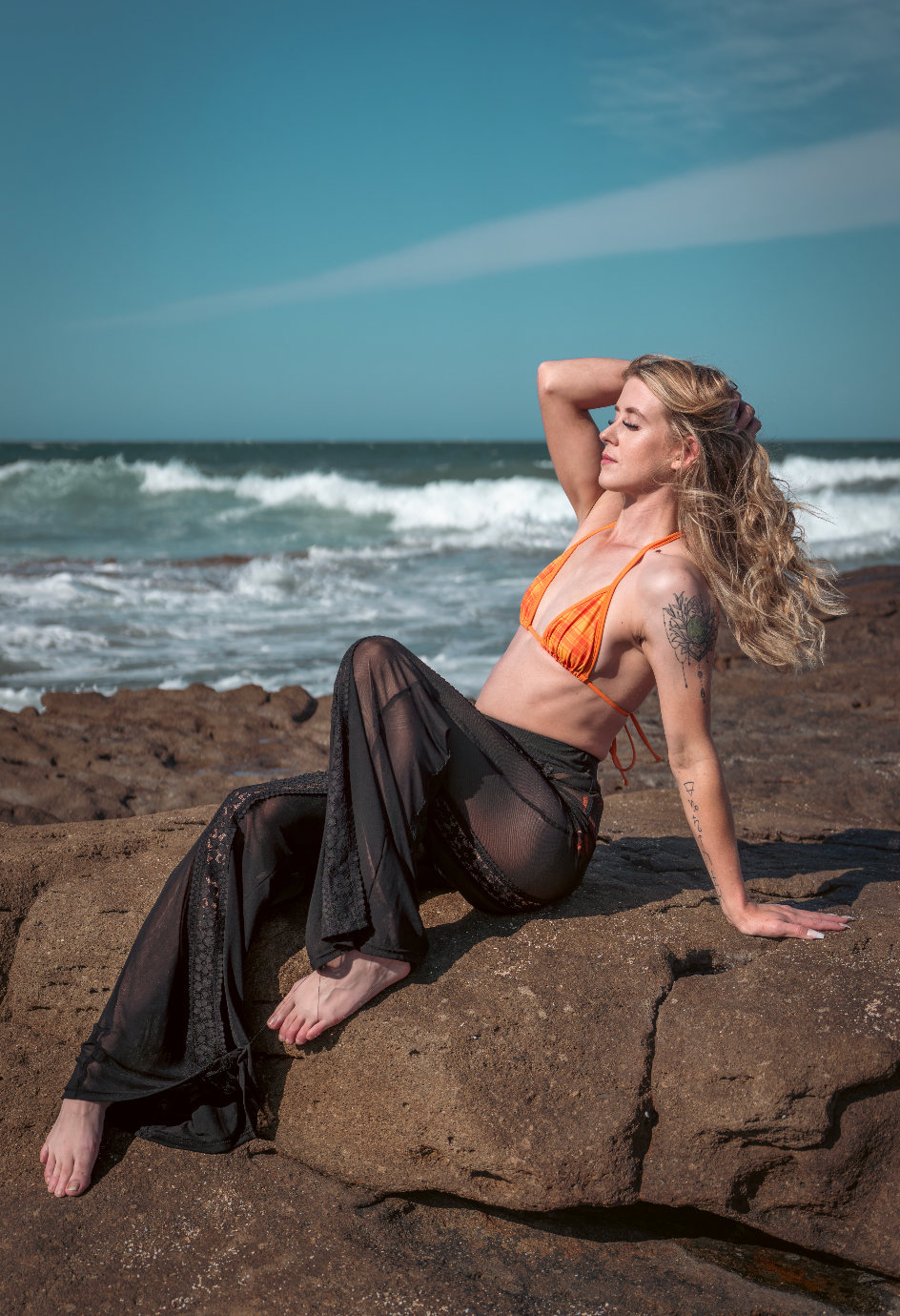 Model in flowing black dress posing dramatically on coastal rocks with ocean waves in background.