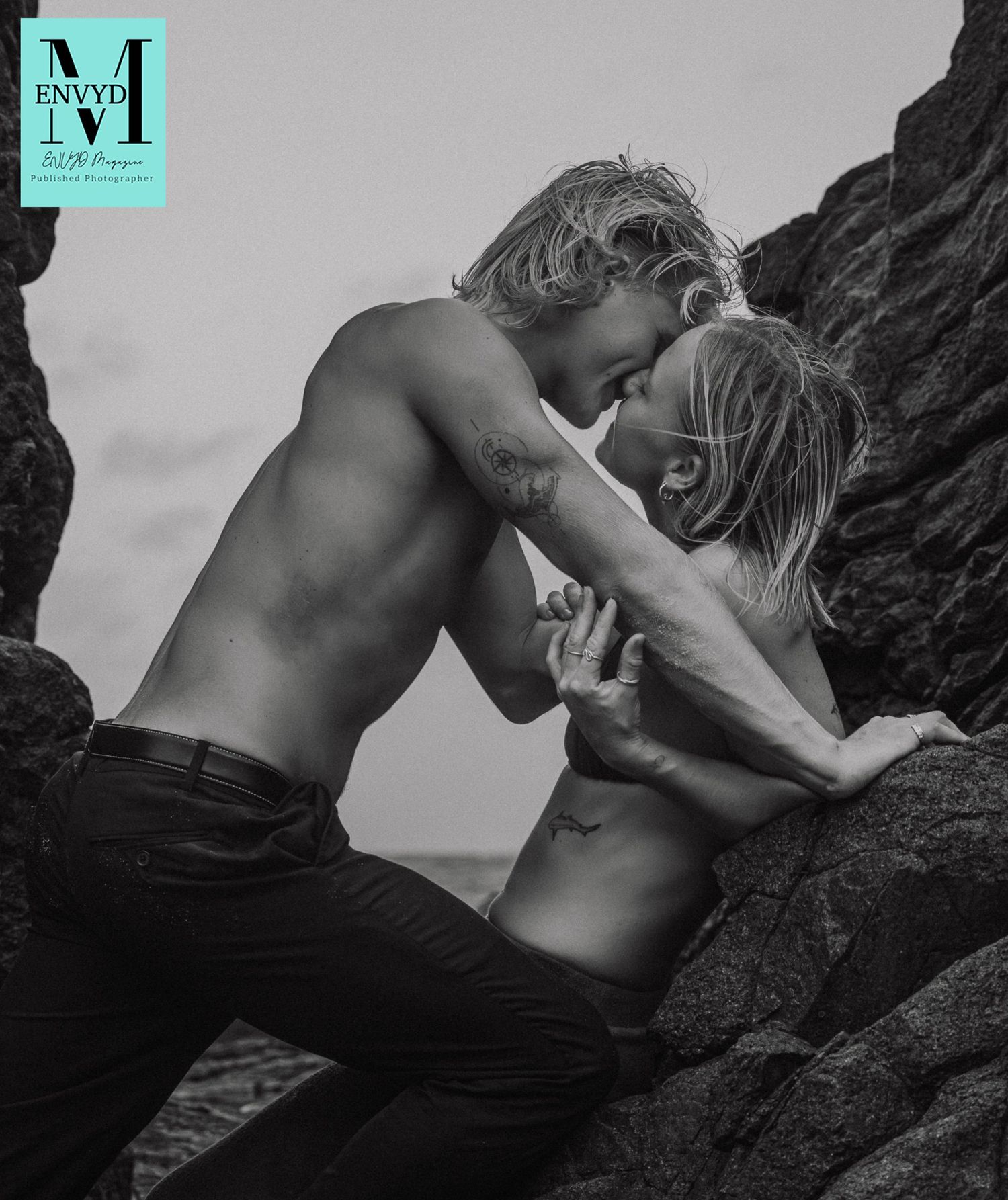 Black and white photo of a shirtless couple kissing romantically among rocky cliffs by the ocean.