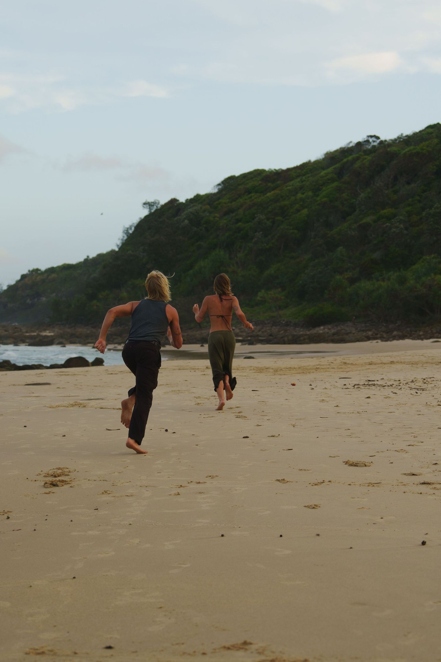 People walking along a scenic sandy beach with lush green hills in the background.