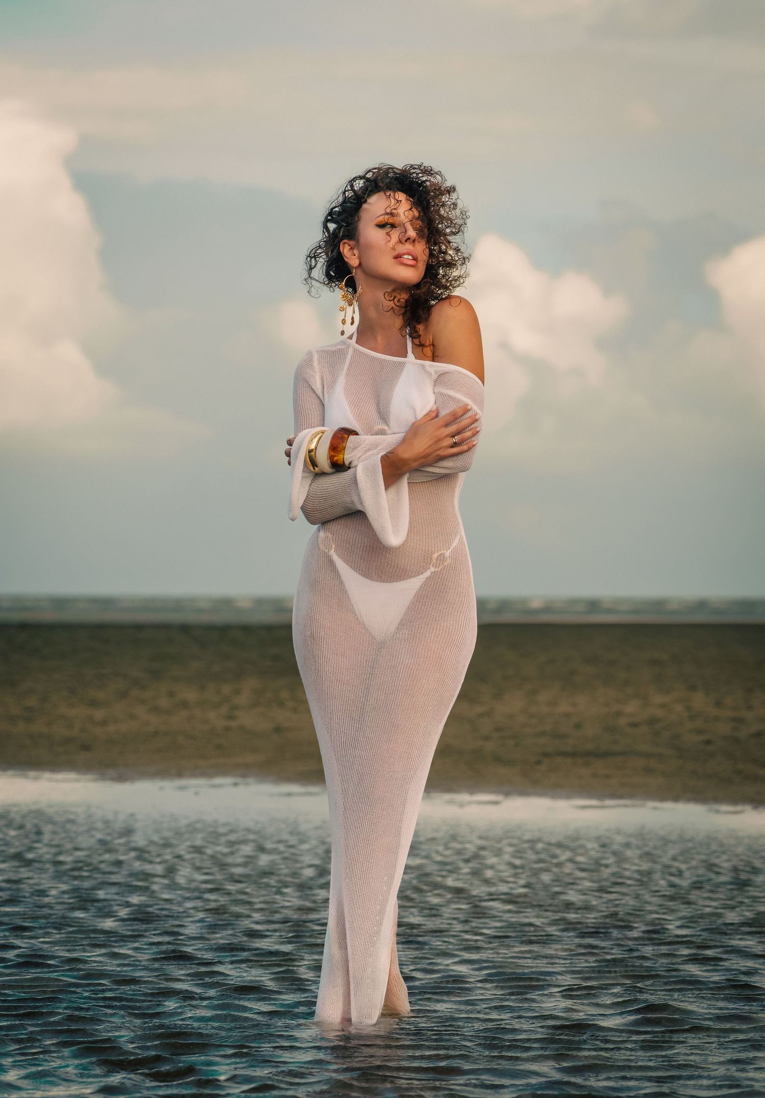 Woman in sheer white gown standing in shallow ocean water with curly hair and gold jewelry.