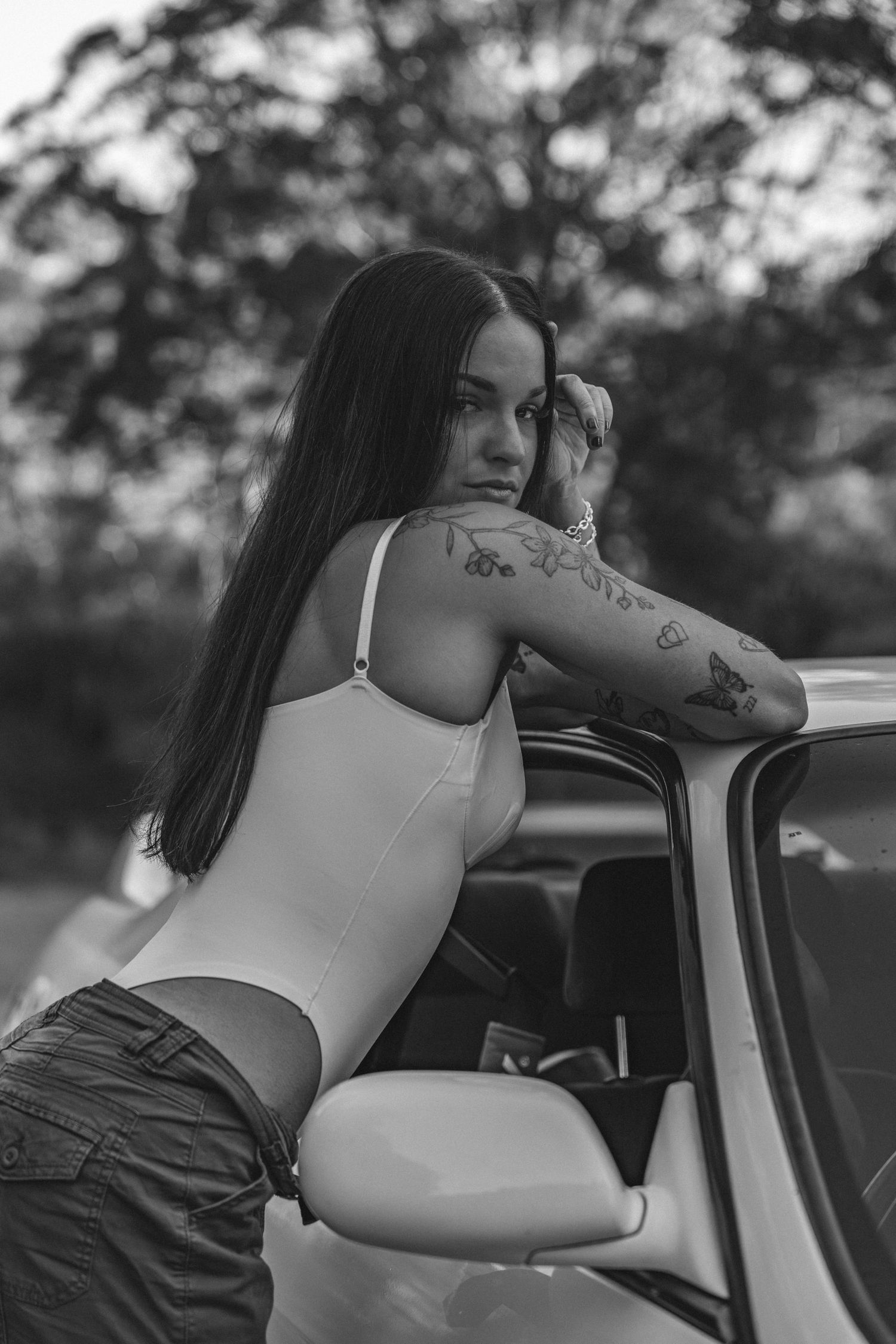 Black and white photo of a tattooed woman leaning on a car door outdoors.