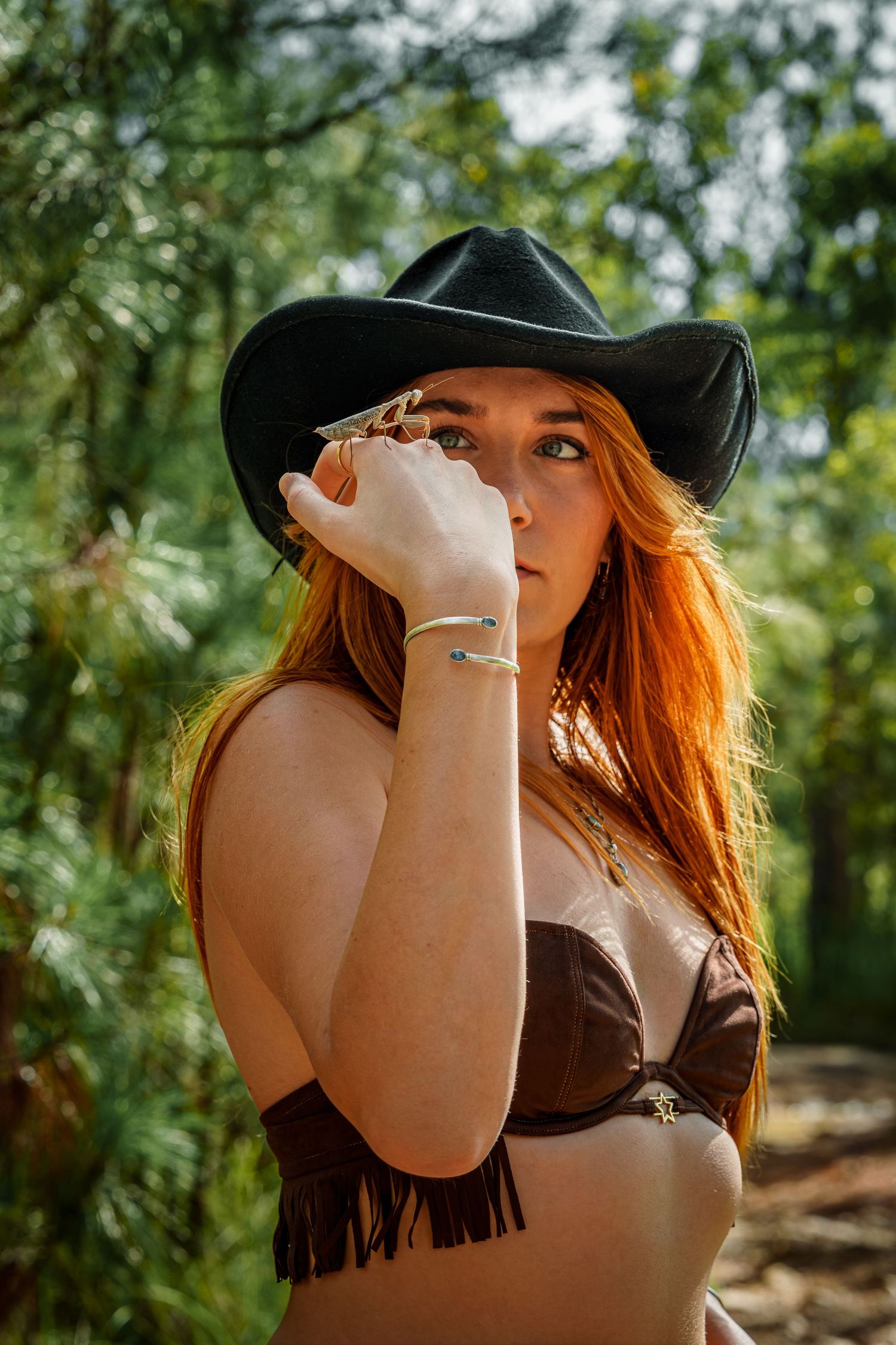A person in a black cowboy hat and brown fringe top poses thoughtfully in a sunlit forest setting.