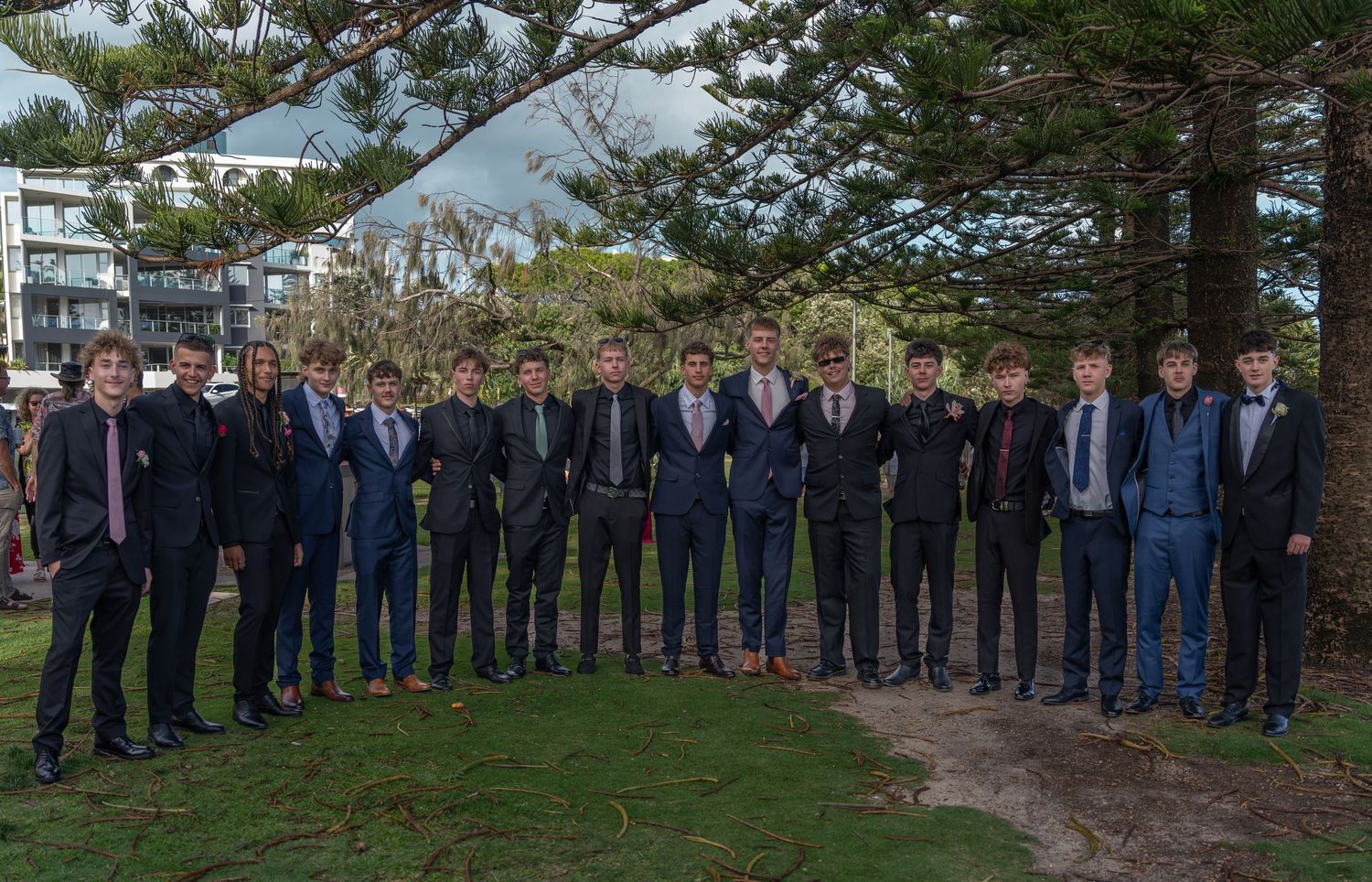 A large group of formally dressed people lined up outdoors near pine trees.