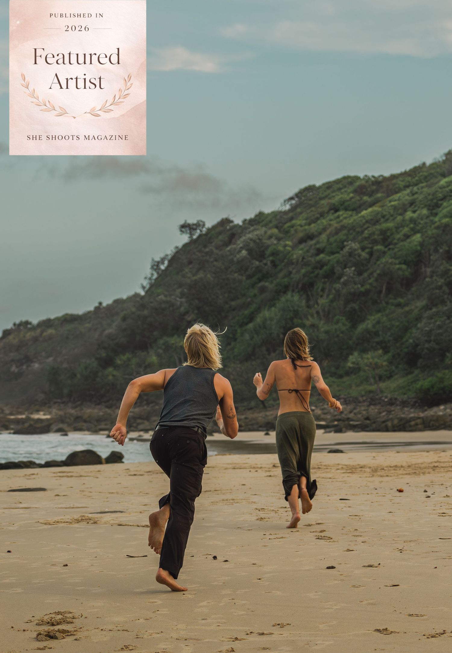 Two people running barefoot on a sandy beach toward lush green headland, featured in She Shoots Magazine 2026.