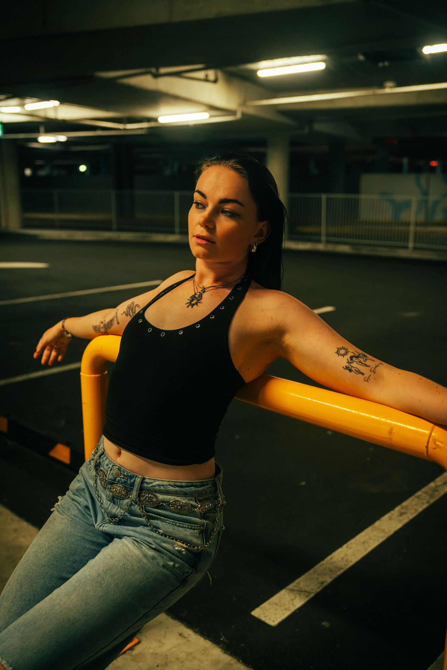 Tattooed woman in black crop top and jeans leaning on yellow railing in dimly lit parking garage.