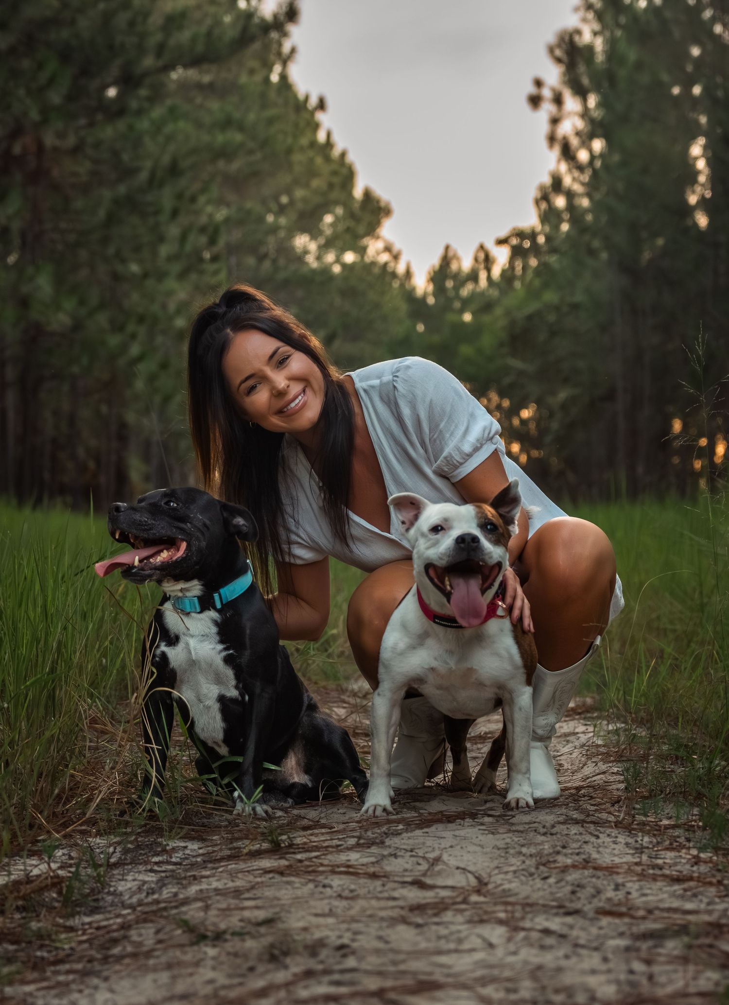 Smiling woman crouching with two dogs on a forest trail at sunset, one black and white dog and one white bulldog.