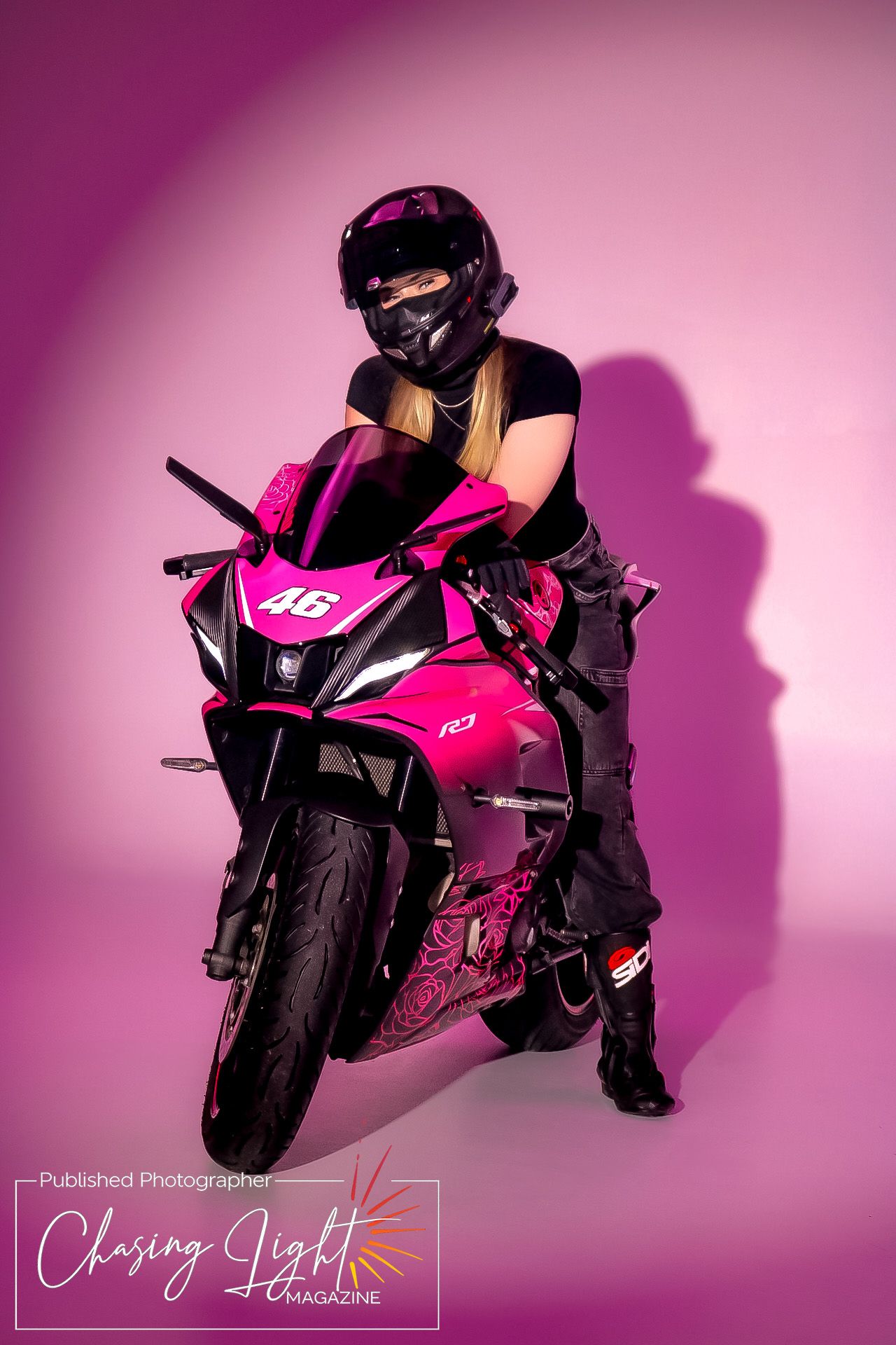 Woman in black helmet poses with pink Yamaha R3 motorcycle numbered 46 against pink studio background.