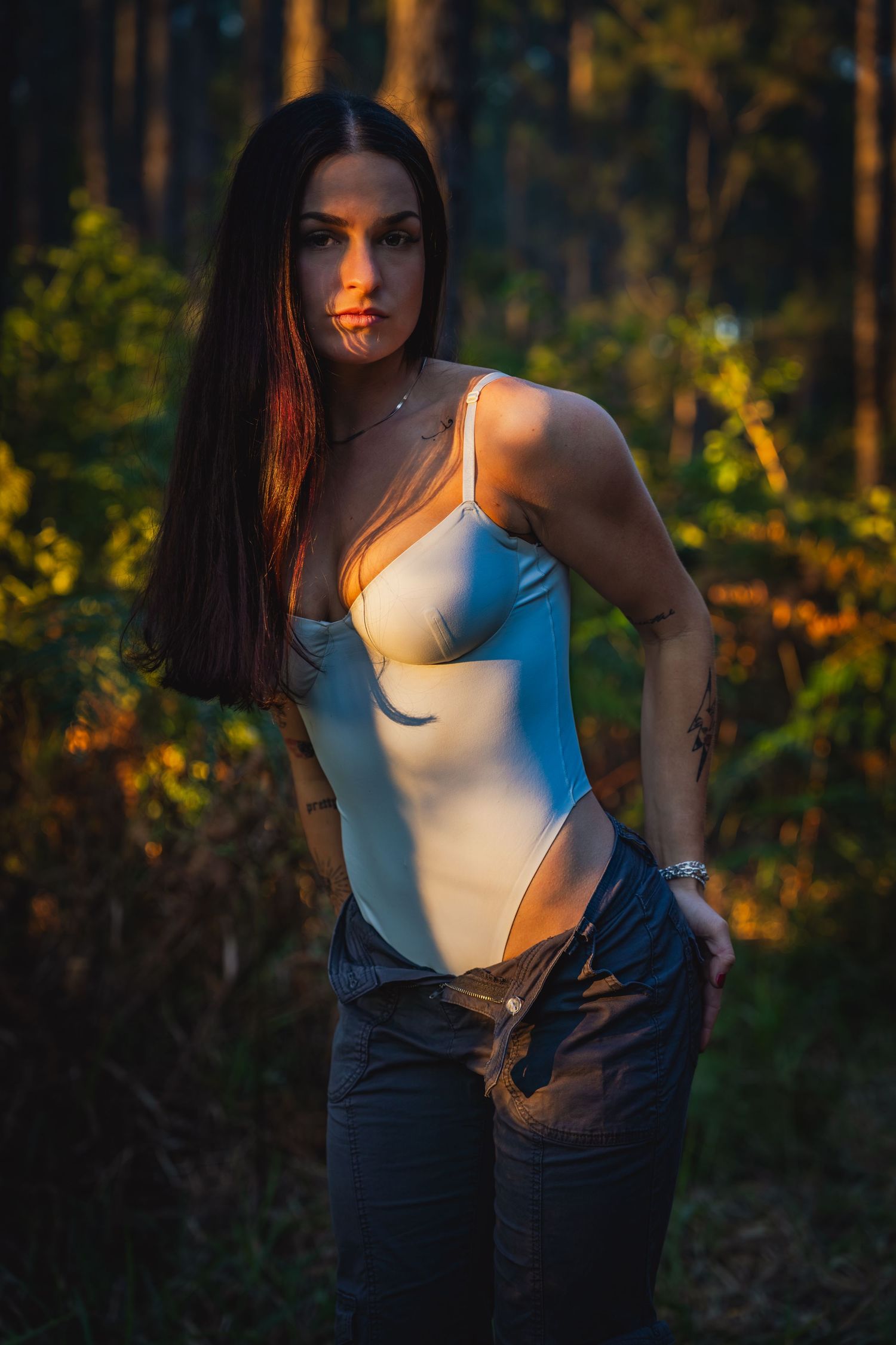 Sunset portrait in white bodysuit and jeans against forest backdrop.