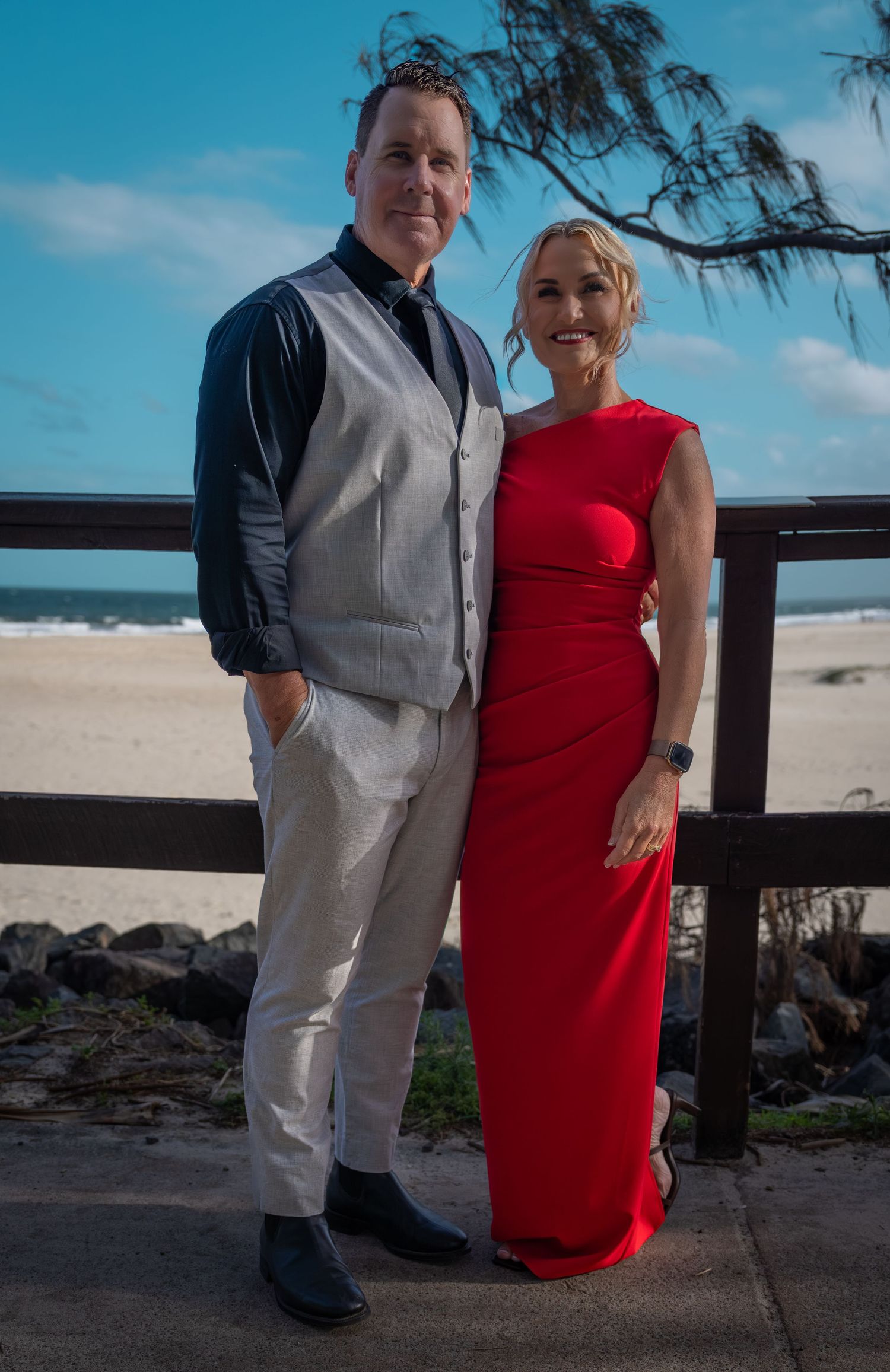 A couple poses together on a beach boardwalk, with the woman wearing a red dress and the man in a light gray suit.