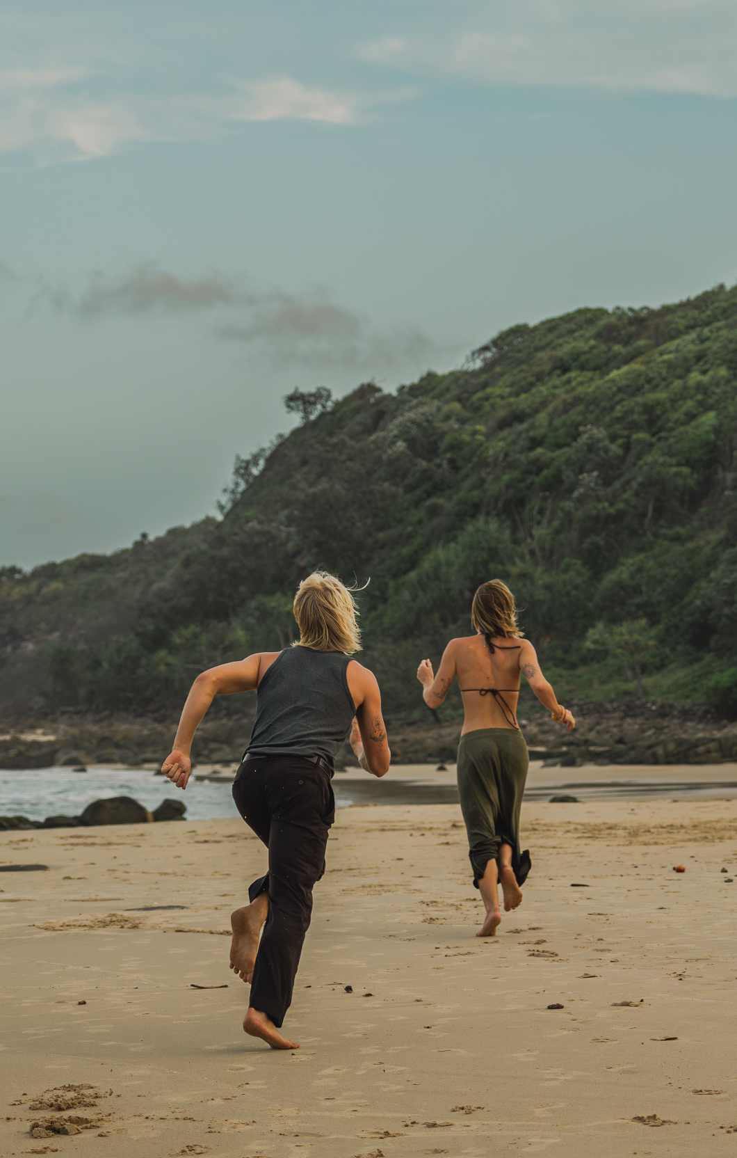 Two people running barefoot on a sandy beach toward lush green headland under a cloudy sky.