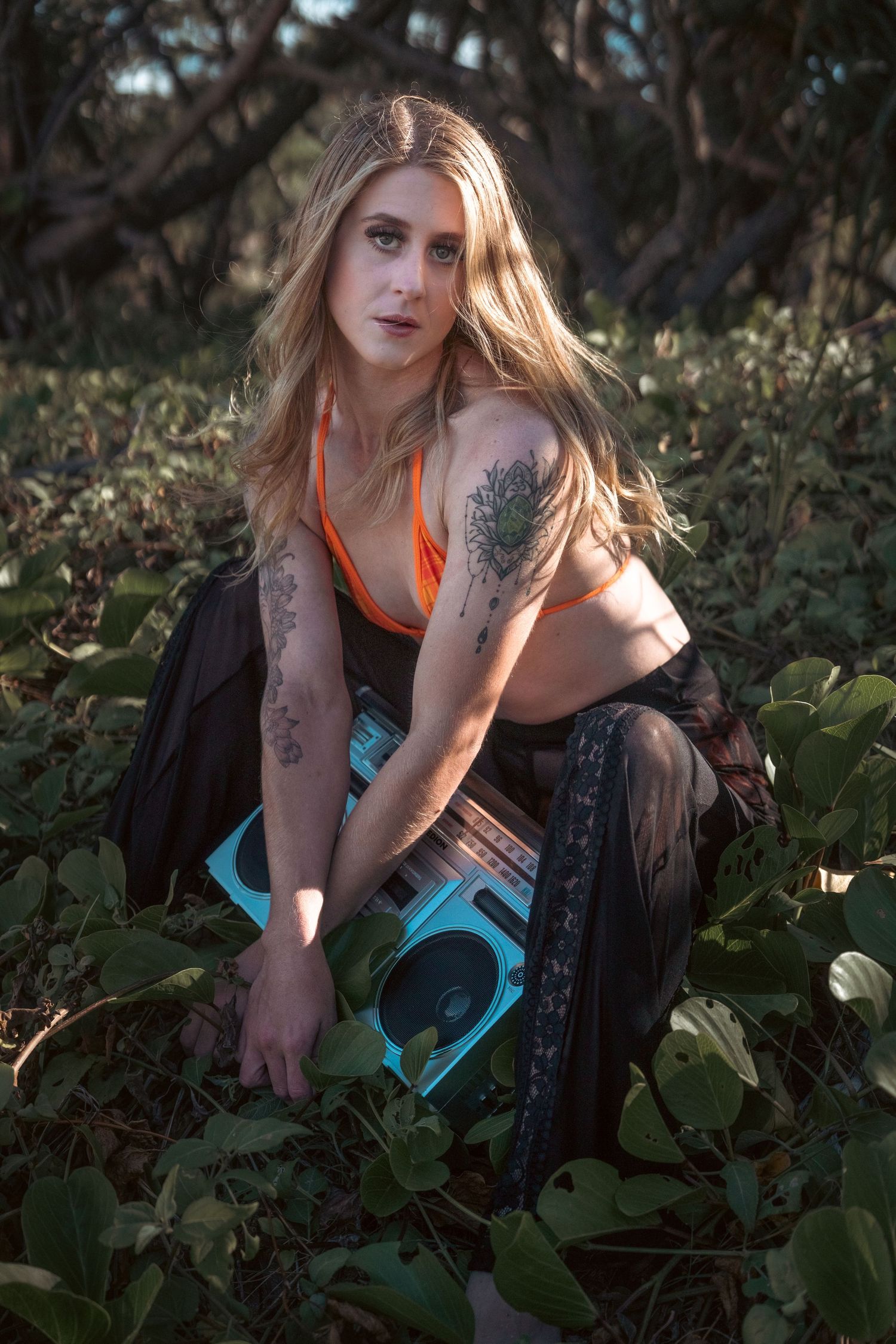 Model in orange top poses with skateboard among foliage in natural lighting.